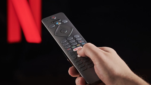 Detail of a mans hand pressing the Netflix button on a TV remote control, with Netflix streaming on a television in the background, taken on March 6, 2020.