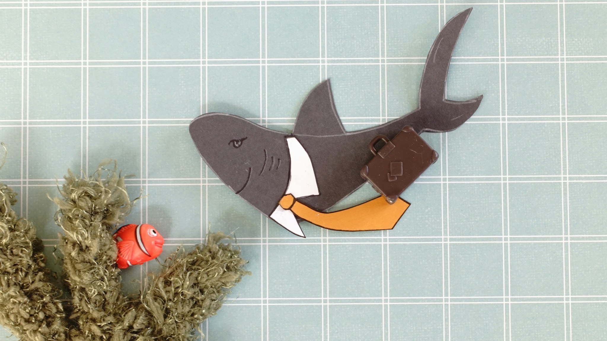 Merged mediums. Loan shark. Paper illustration of a shark in a white collar and tie holding a plastic case looking down on a small clown fish looking out of knitted seaweed.