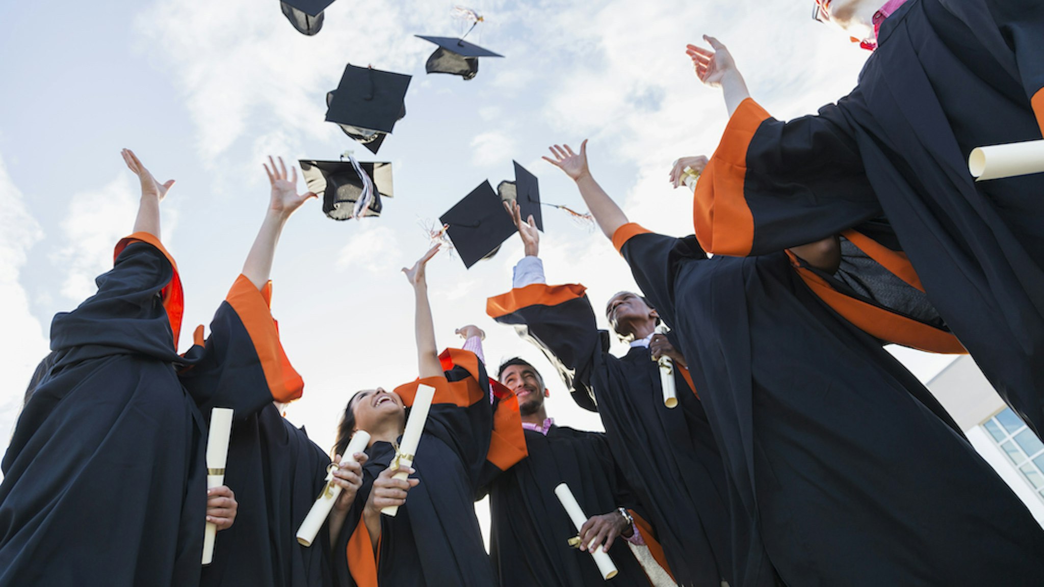 A group of seven multi-ethnic high school or university graduates wearing graduation gowns, holding diplomas. Throwing their caps in the air.