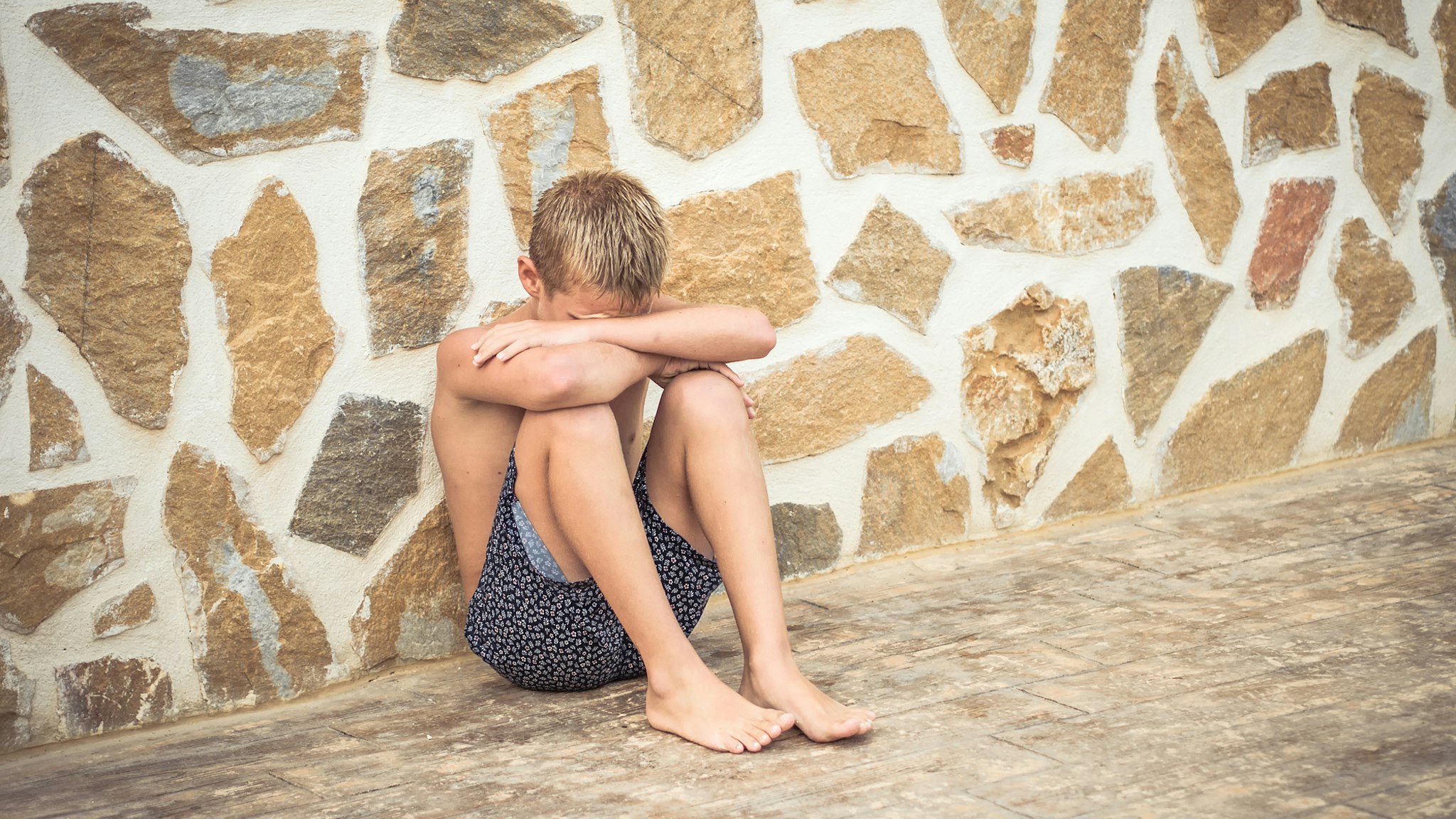 Displeased teenager dressed in swimsuit crouching against the wall and hiding his head.