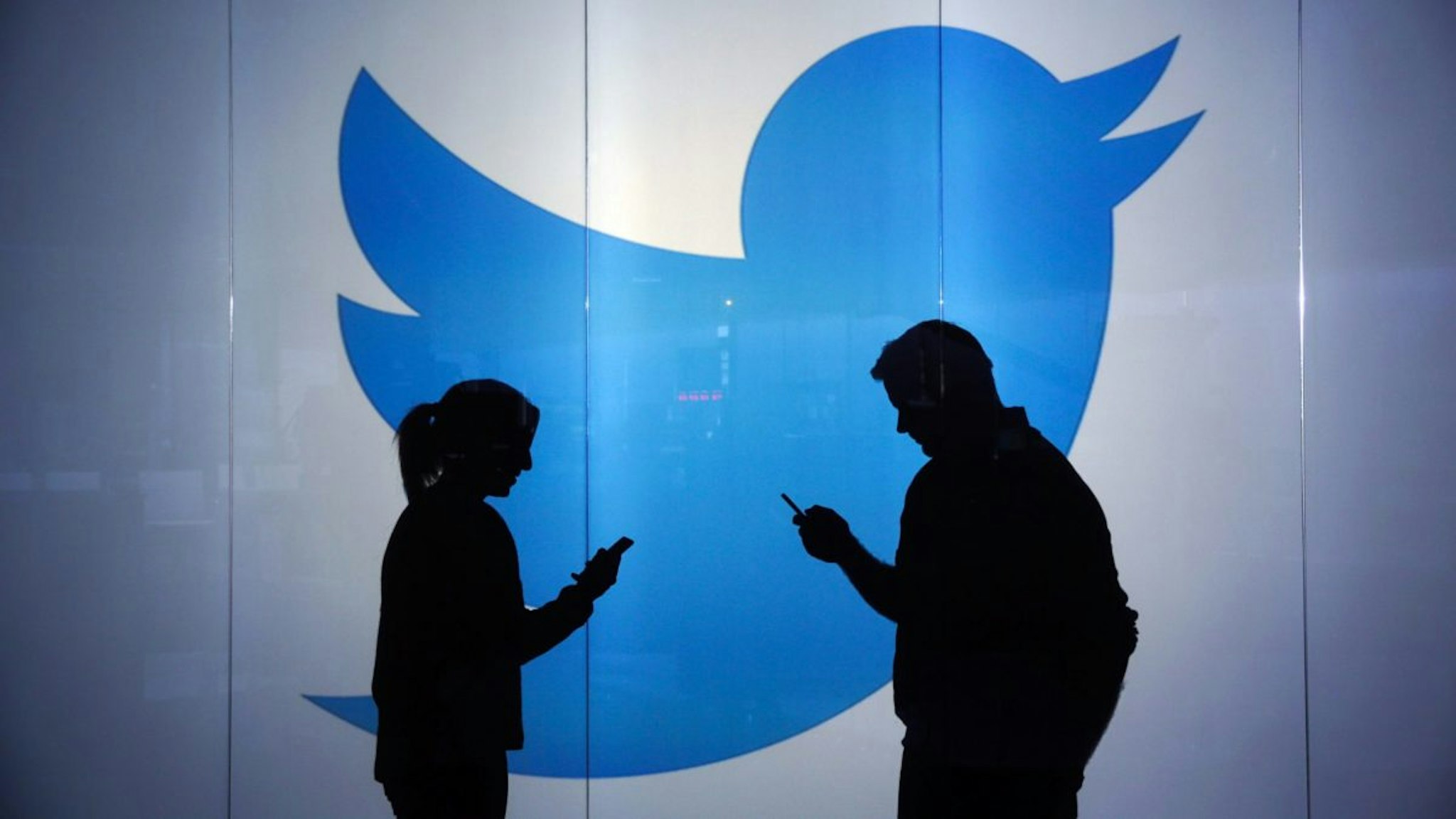 People are seen as silhouettes as they check mobile devices whilst standing against an illuminated wall bearing Twitter Inc.'s logo in this arranged photograph in London, U.K., on Tuesday, Jan. 5, 2016.