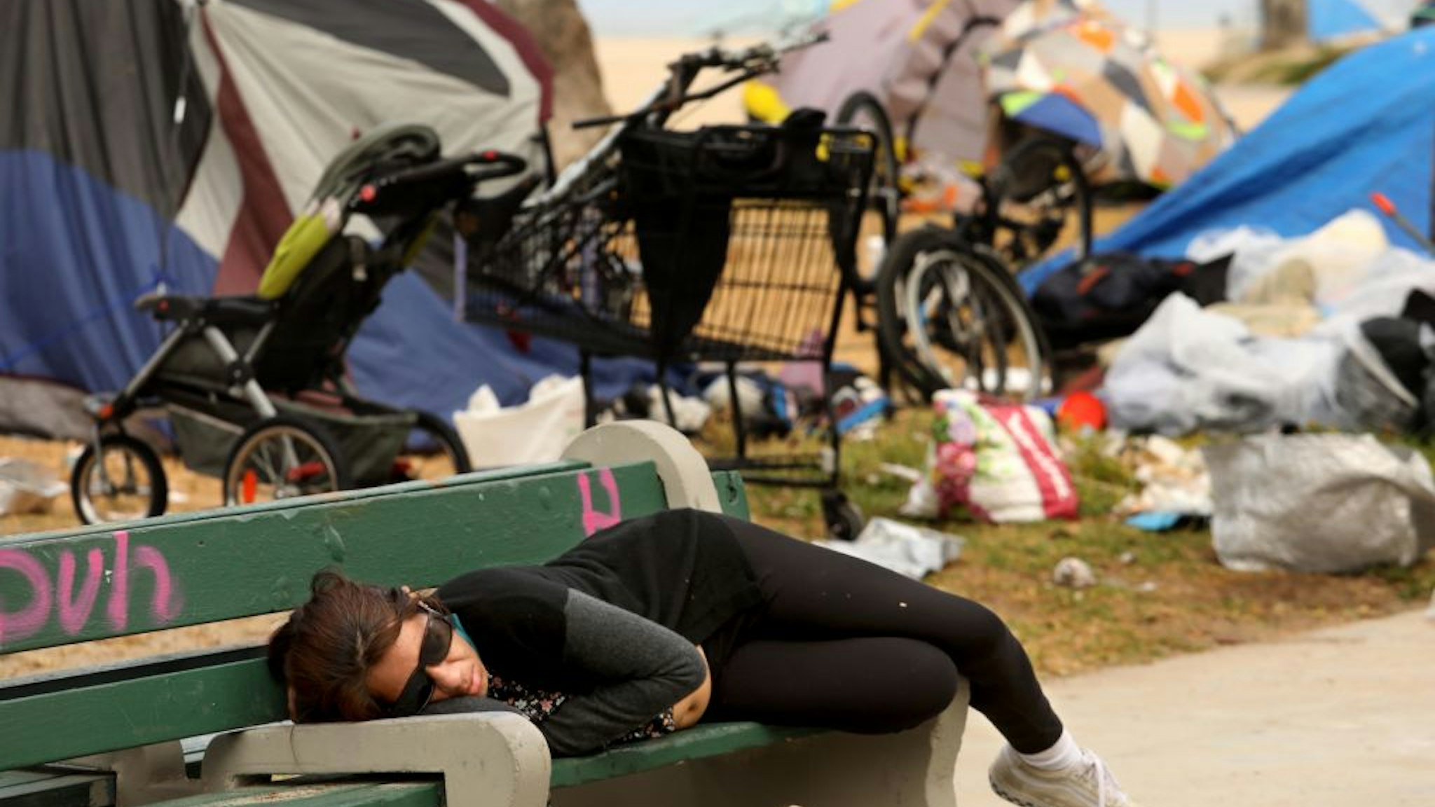 VENICE, CA - MAY 18, 2021 - - A woman sleeps on a bench against a backdrop of homeless encampments along Ocean Front Walk in Venice May 18, 2021.