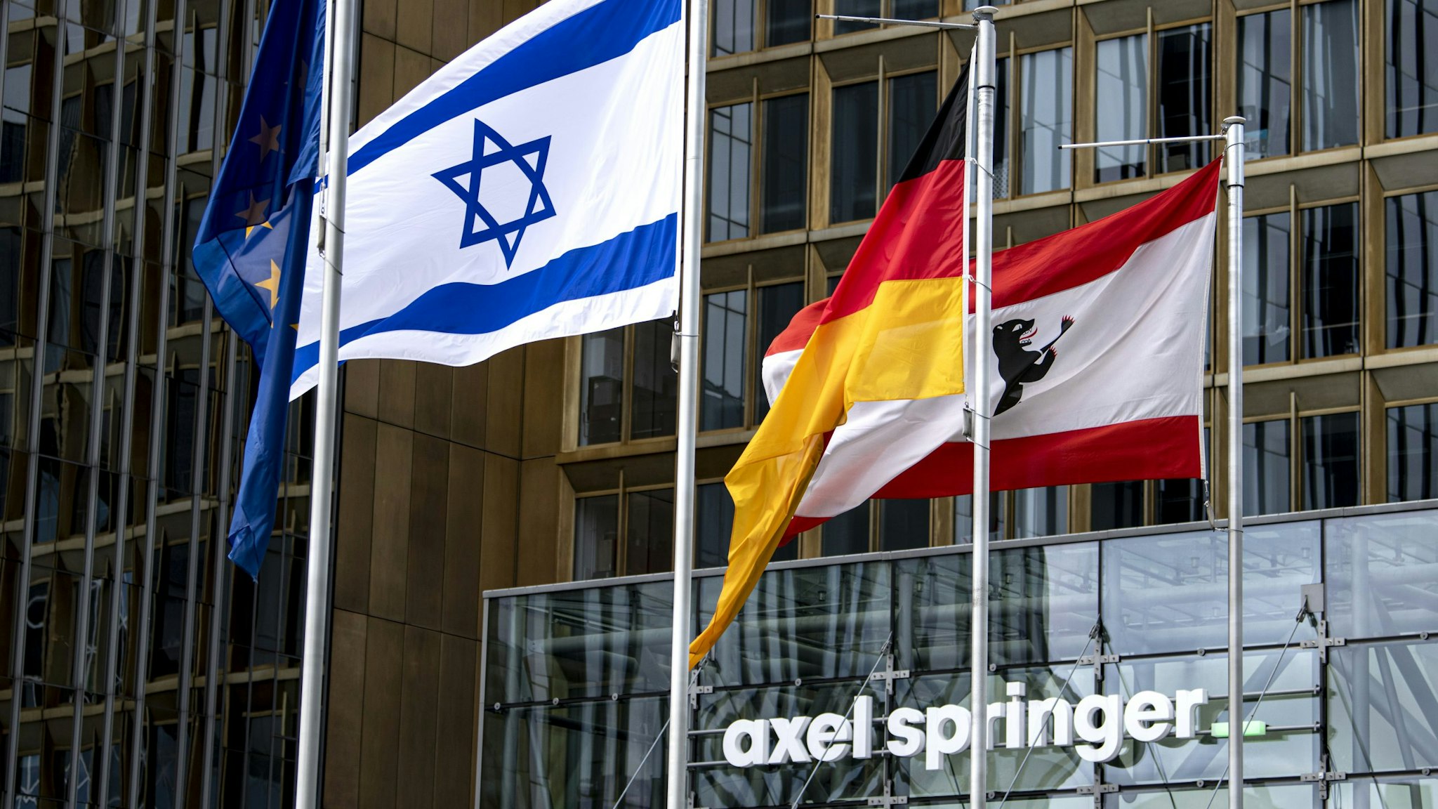 16 May 2021, Berlin: The Israeli flag, which was raised on Sunday morning, flies in front of the Axel Springer building. In addition, the (l-r) European, German and Berlin flags can be seen.