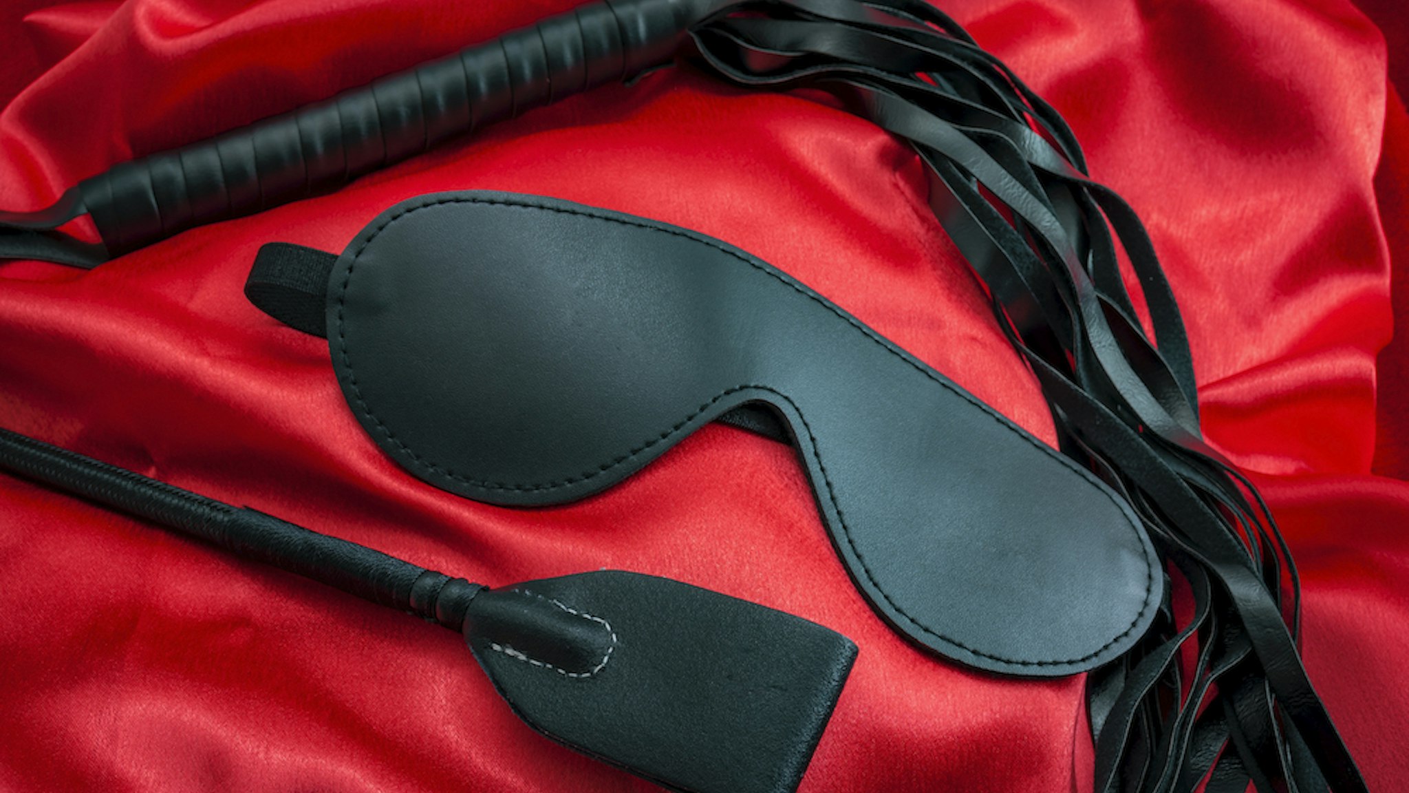 Riding crop, a whip flogger and blindfold mask on red satin, kinky sex toys for dom. (Moussa81 via Getty Images)
