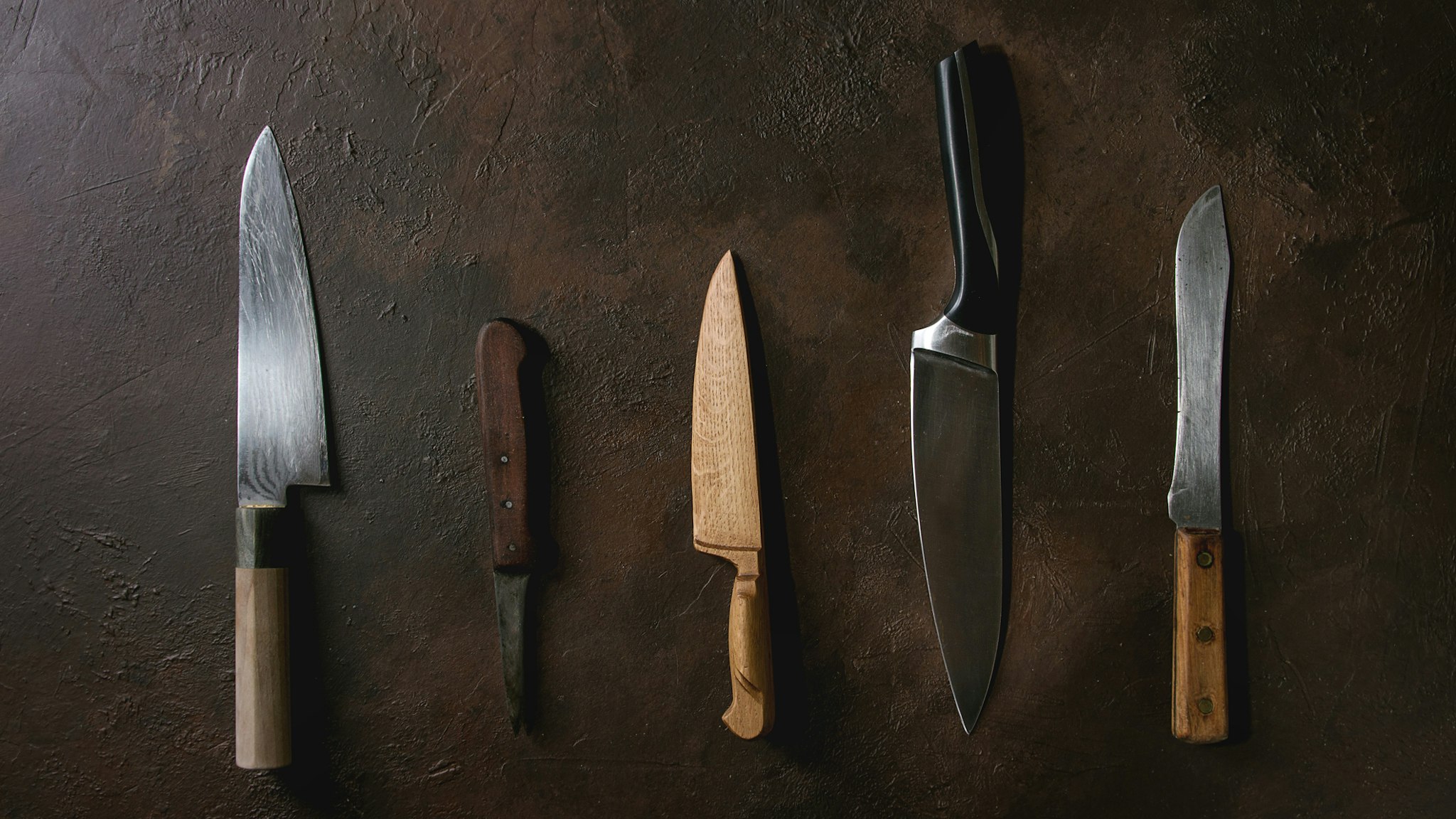 Collection of different kitchen metal, wooden and vintage knives in row over dark brown texture background. Flat lay, space