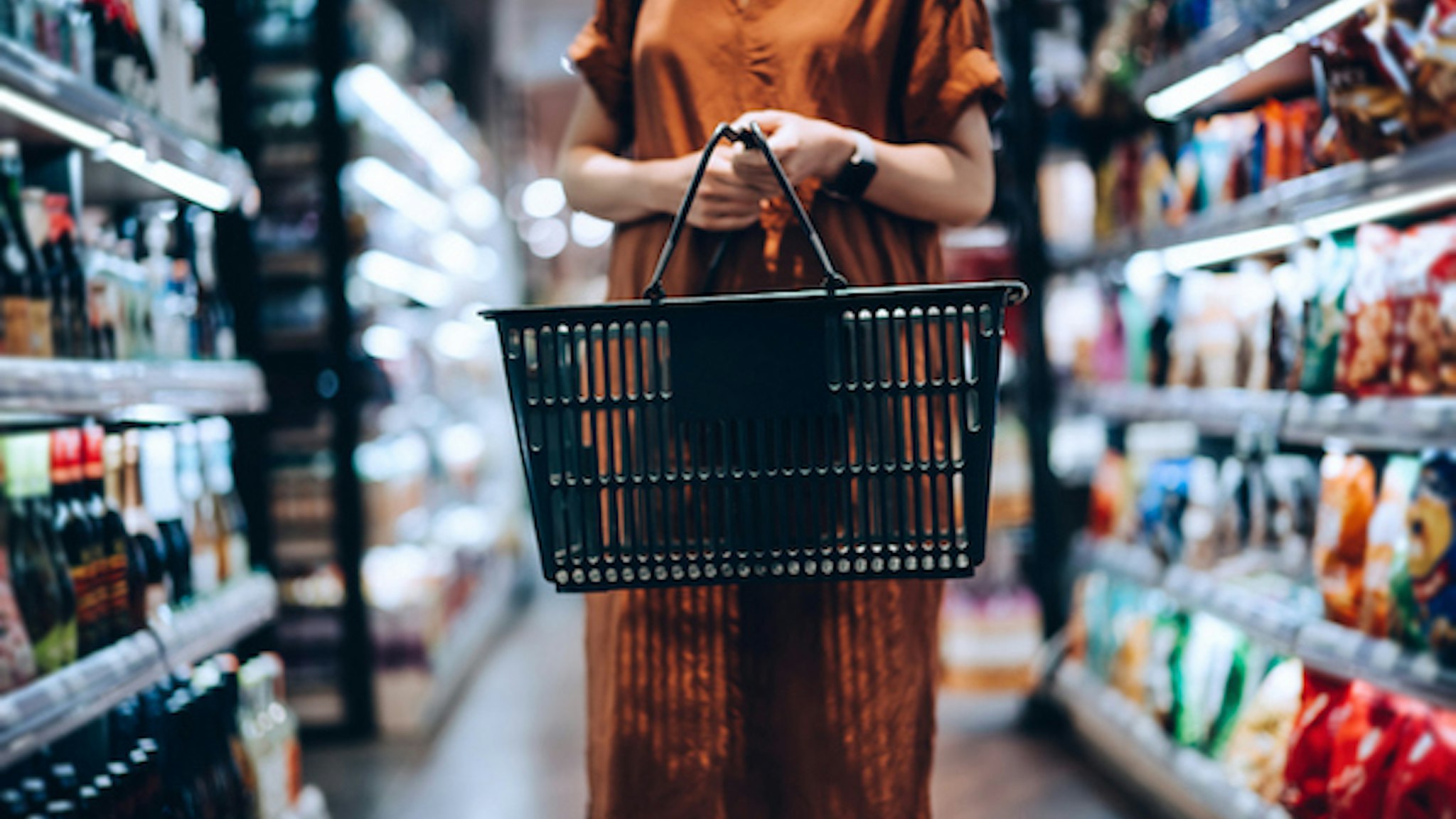 Cropped shot of young woman carrying a shopping basket, standing along the product aisle, grocery shopping for daily necessities in supermarket - stock photo Cropped shot of young woman carrying a shopping basket, standing along the product aisle, grocery shopping for daily necessities in supermarket d3sign via Getty Images