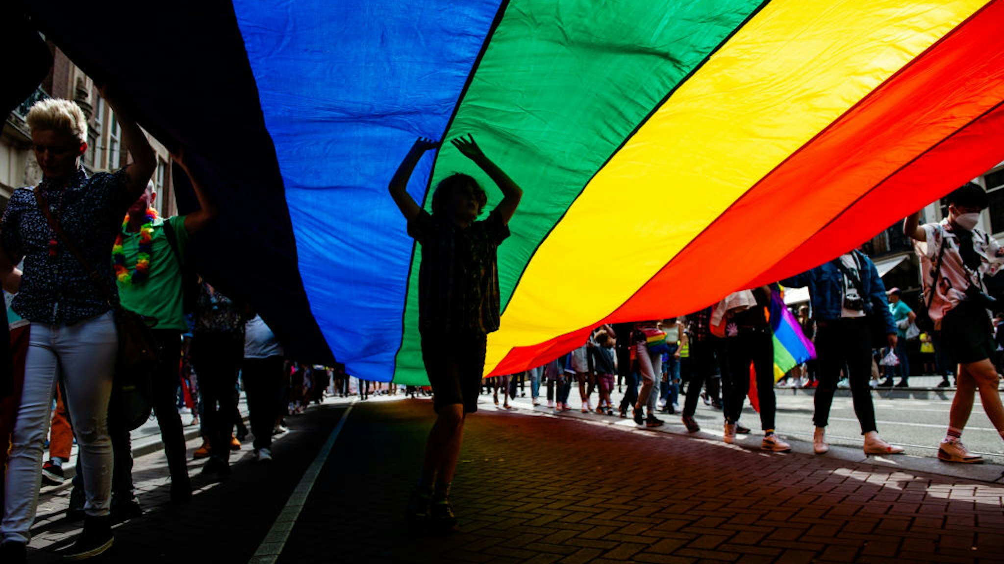 A boy is dancing under the big rainbow flag, during the celebration of the Pride walk in Amsterdam, on August 7th, 2021.