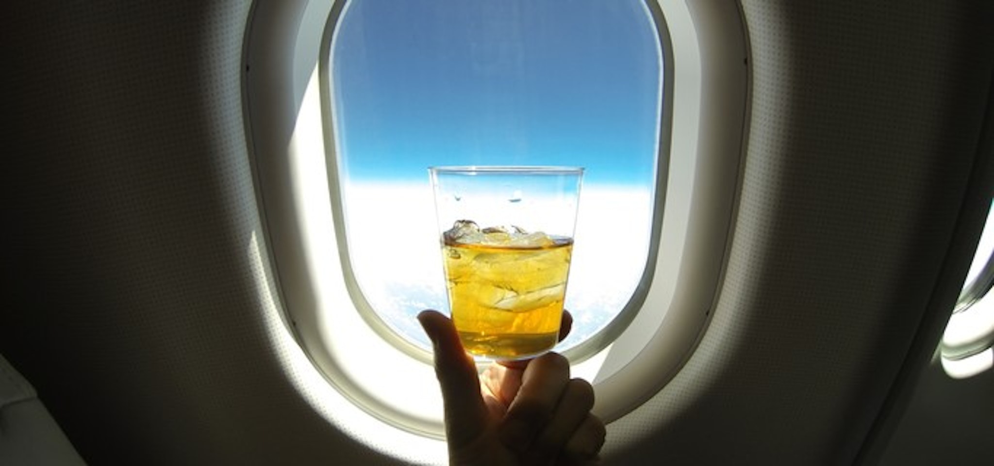 Refreshing glass of ice tea (or alcohol mix) centered on the window of a jumbo airplane, with blue sky above and snowy white clowds below.
