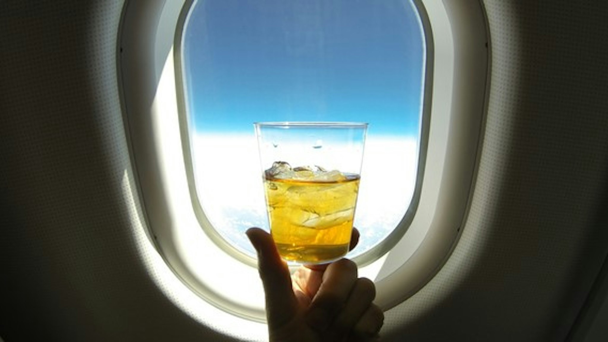 Refreshing glass of ice tea (or alcohol mix) centered on the window of a jumbo airplane, with blue sky above and snowy white clowds below.
