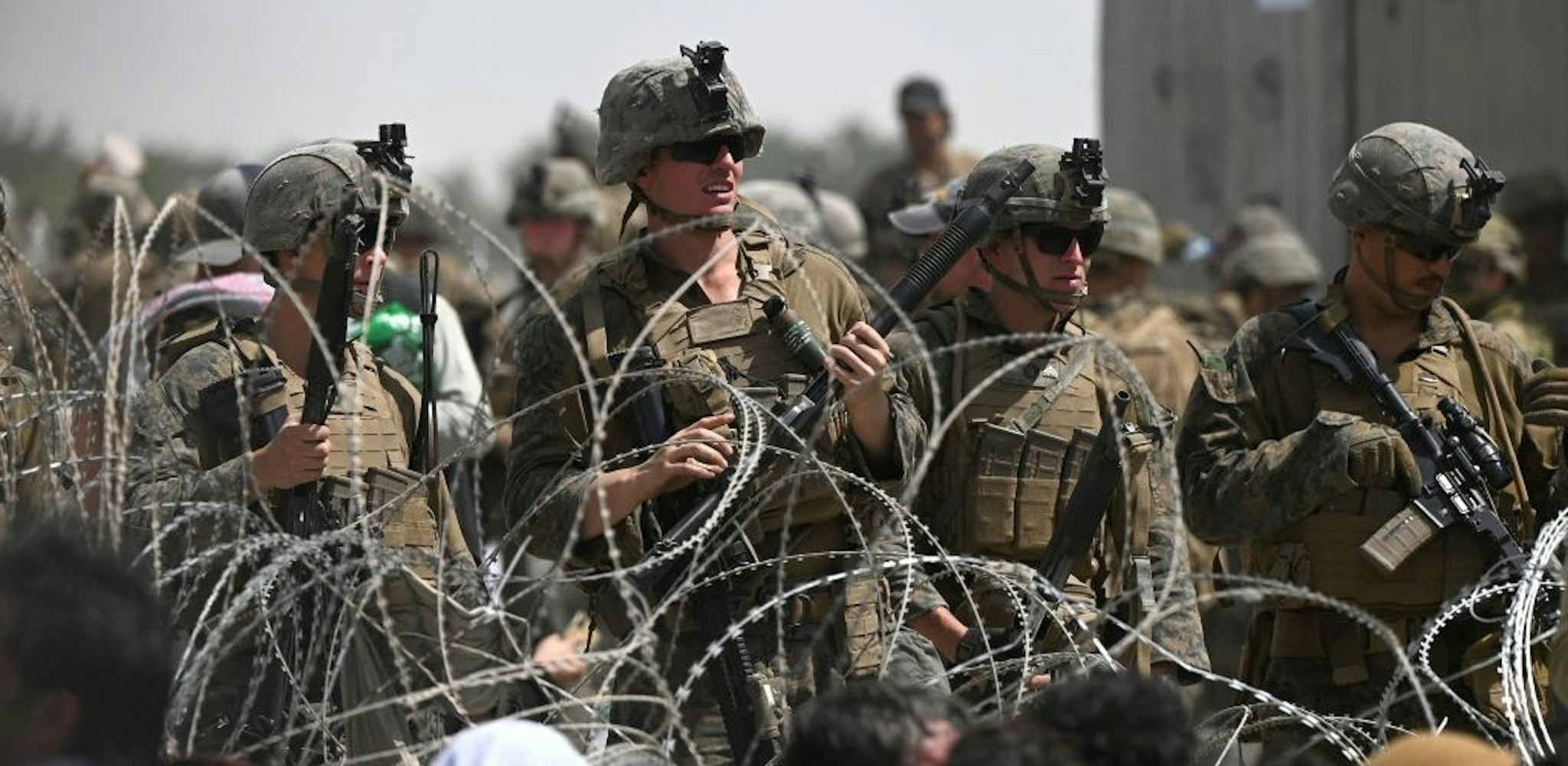 TOPSHOT - US soldiers stand guard behind barbed wire as Afghans sit on a roadside near the military part of the airport in Kabul on August 20, 2021, hoping to flee from the country after the Taliban's military takeover of Afghanistan. (Photo by Wakil KOHSAR / AFP) (Photo by