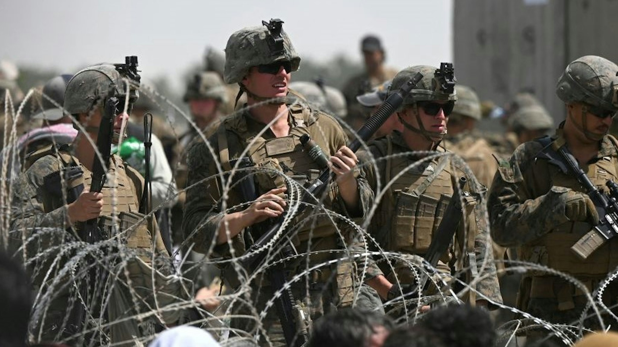 TOPSHOT - US soldiers stand guard behind barbed wire as Afghans sit on a roadside near the military part of the airport in Kabul on August 20, 2021, hoping to flee from the country after the Taliban's military takeover of Afghanistan. (Photo by Wakil KOHSAR / AFP) (Photo by