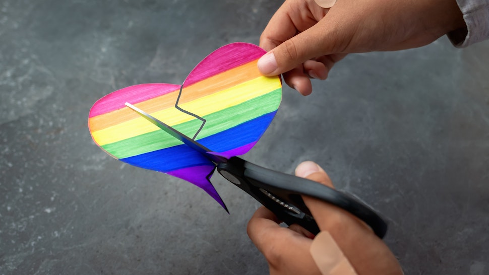 Person with hands covered with medical plasters cuts heart painted in colors of LGBT flag with large scissors.