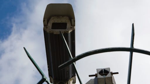 Security camera CCTV camera fixed to a building and back lit against a blue cloudy sky.