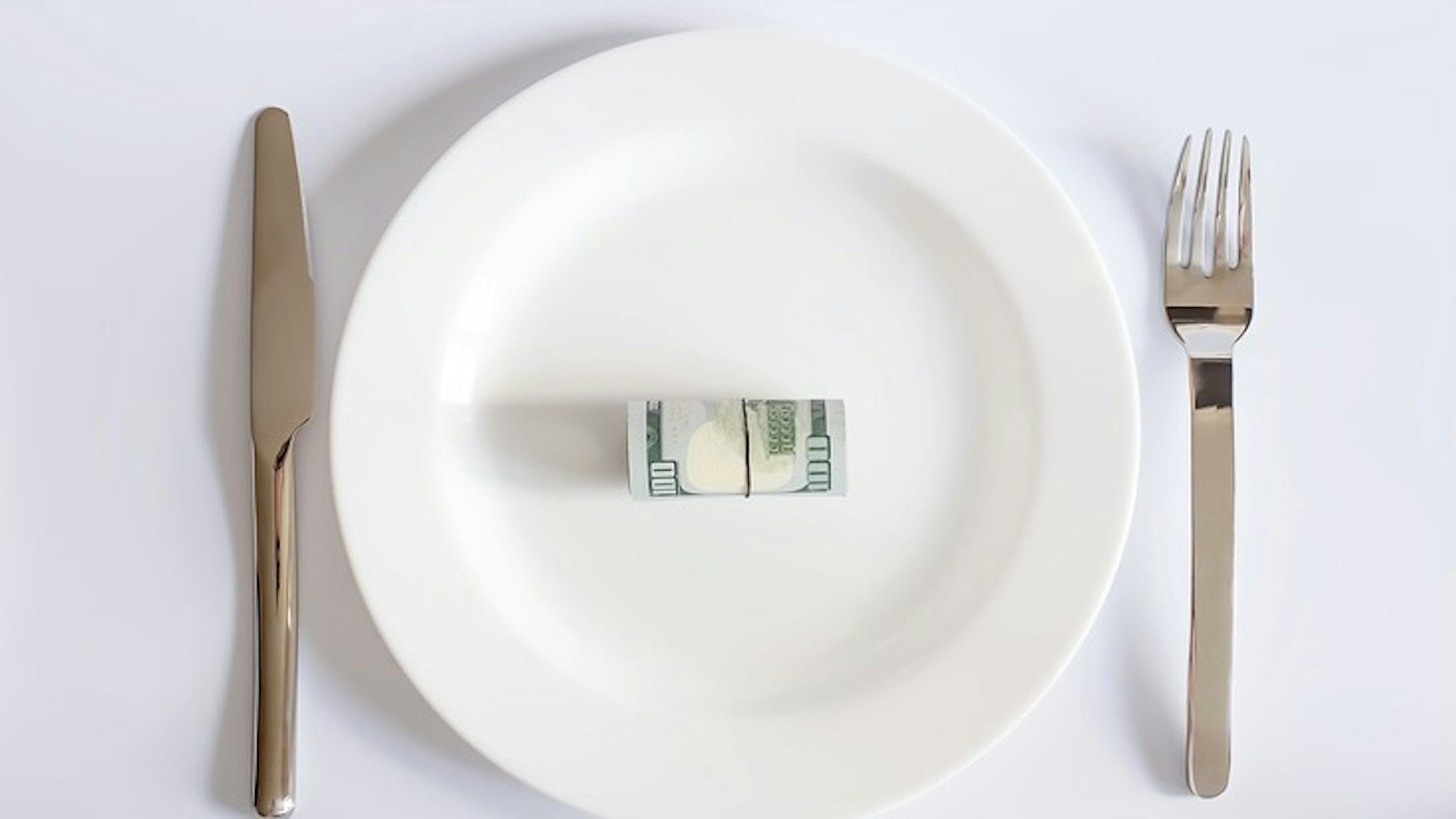 A dollar bill on a white plate between a fork and a knife on white background, - stock photo A dollar bill on a white plate between a fork and a knife on white background,business concept Iryna Drozd via Getty Images
