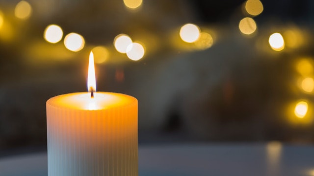 Festive background with burning cylindrical white candle in the foreground and bokeh of sparkling party lights in the background with copy space.