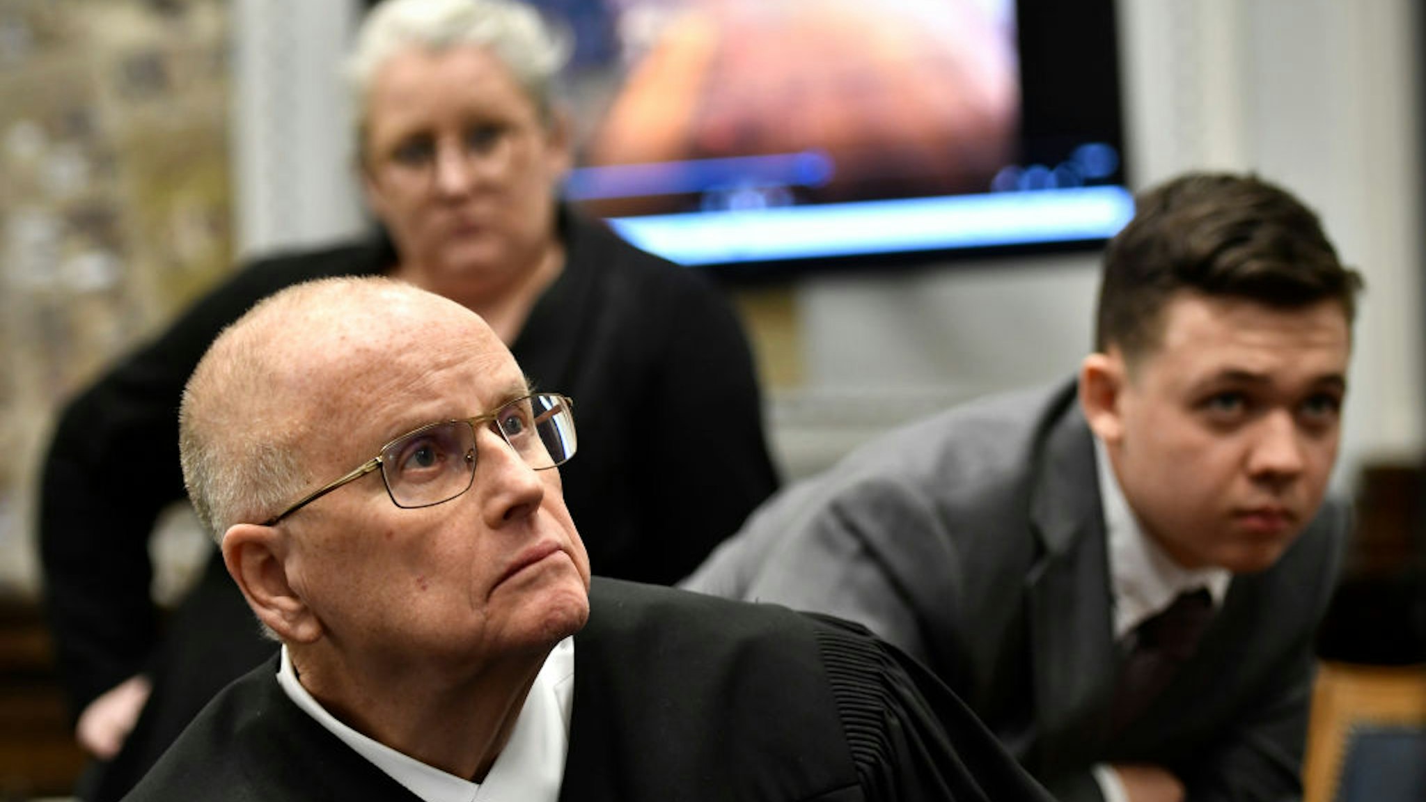 Judge Bruce Schroeder, front, comes down from the bench and sits closer to a 4k television screen to watch a video as Kyle Rittenhouse, right, and his attorney Natalie Wisco stand behind him during proceedings at the Kenosha County Courthouse on November 12, 2021 in Kenosha, Wisconsin.