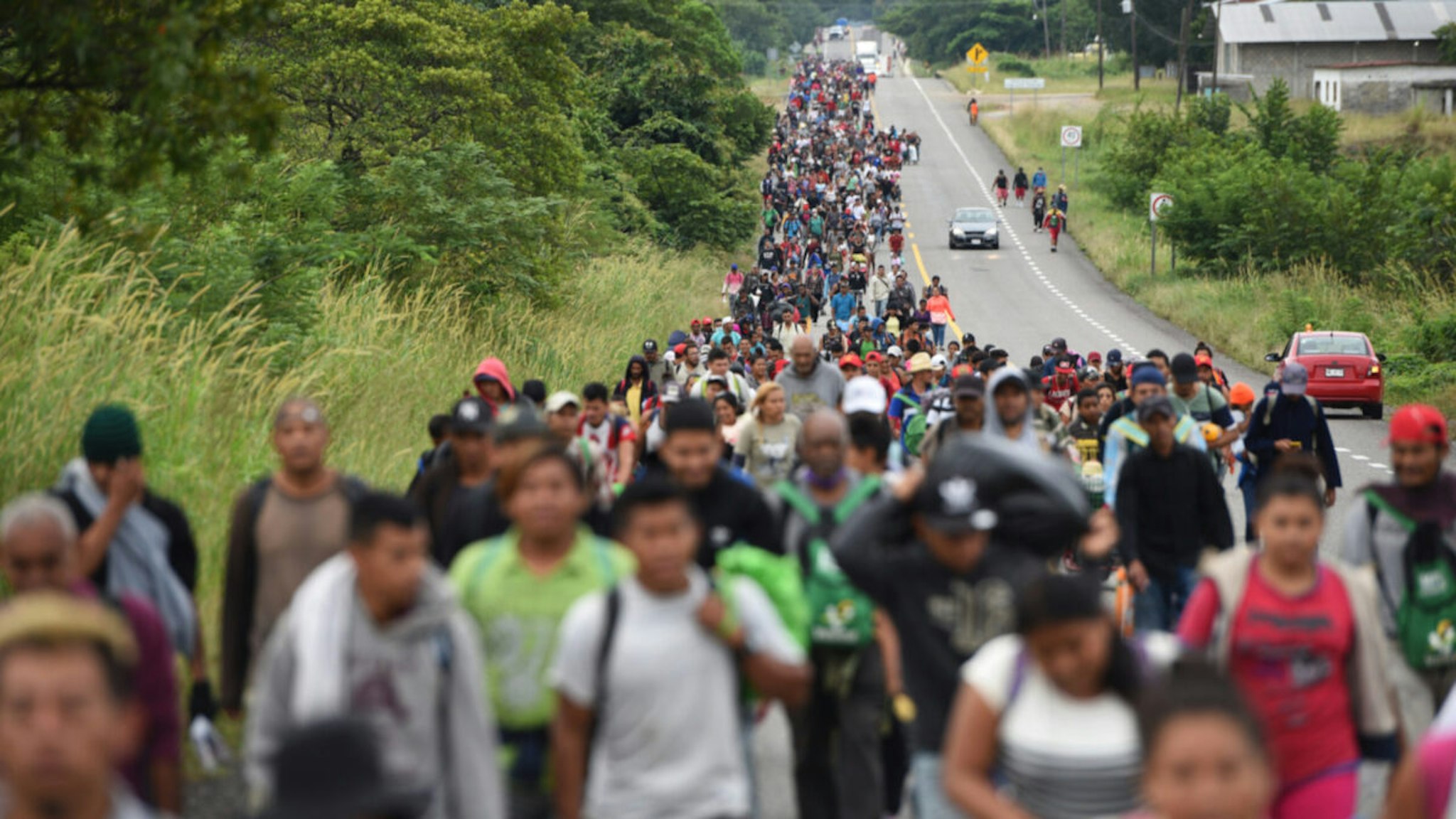 Numerous people from Central America walk together along a rural road towards the US border.