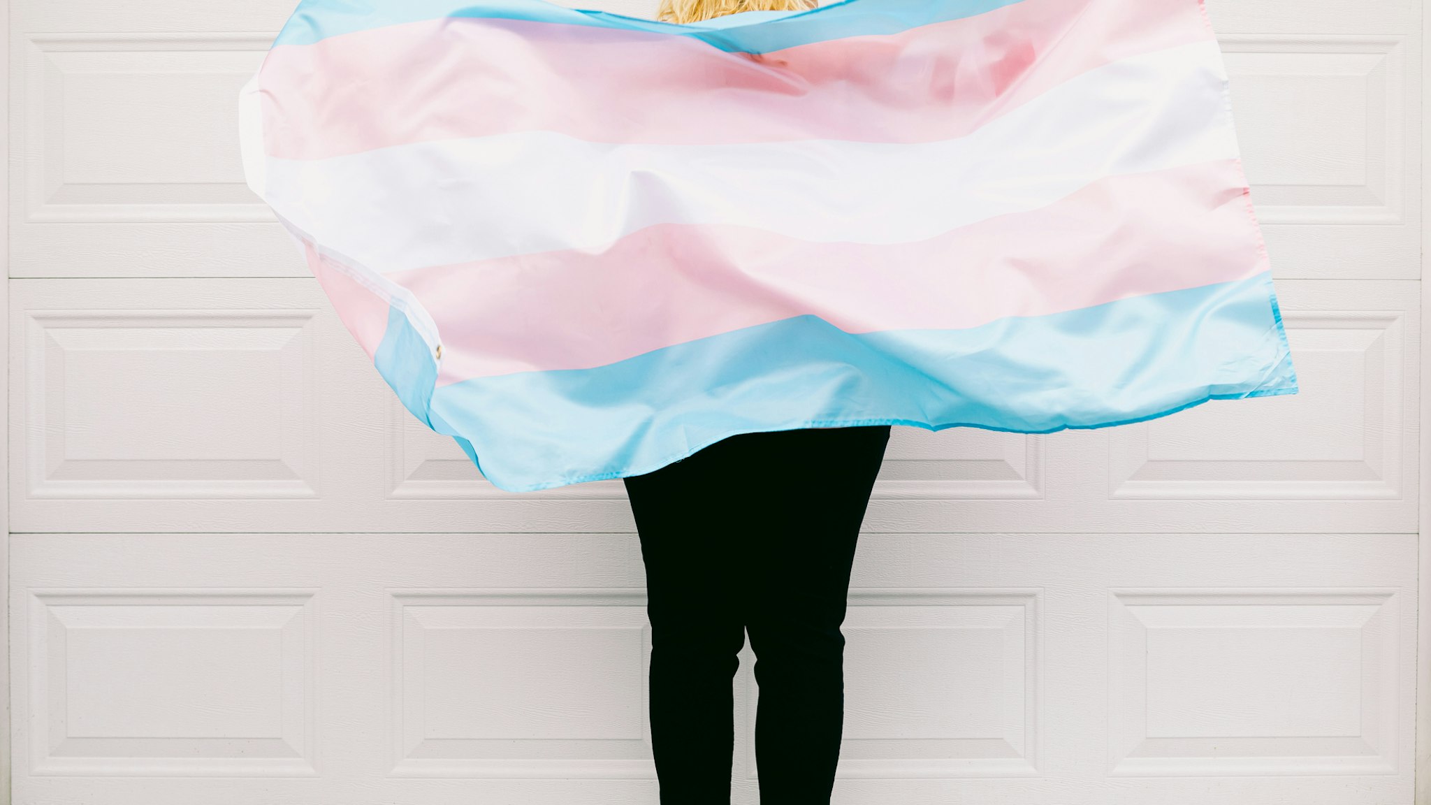 Transgender person from behind, wearing pink and white striped sweatshirt, holds transgender flag - stock photo