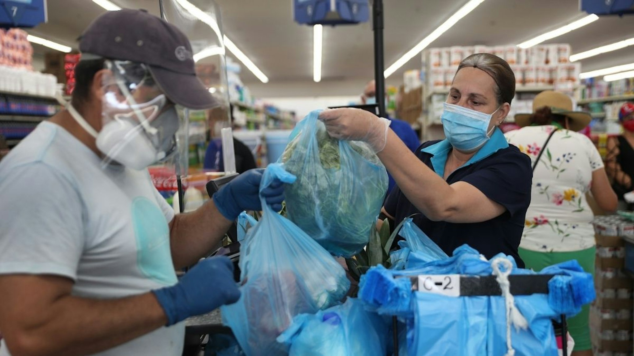 Lay Guzman stands behind a partial protective plastic screen and wears a mask and gloves as she works as a cashier at the Presidente Supermarket on April 13, 2020 in Miami, Florida.