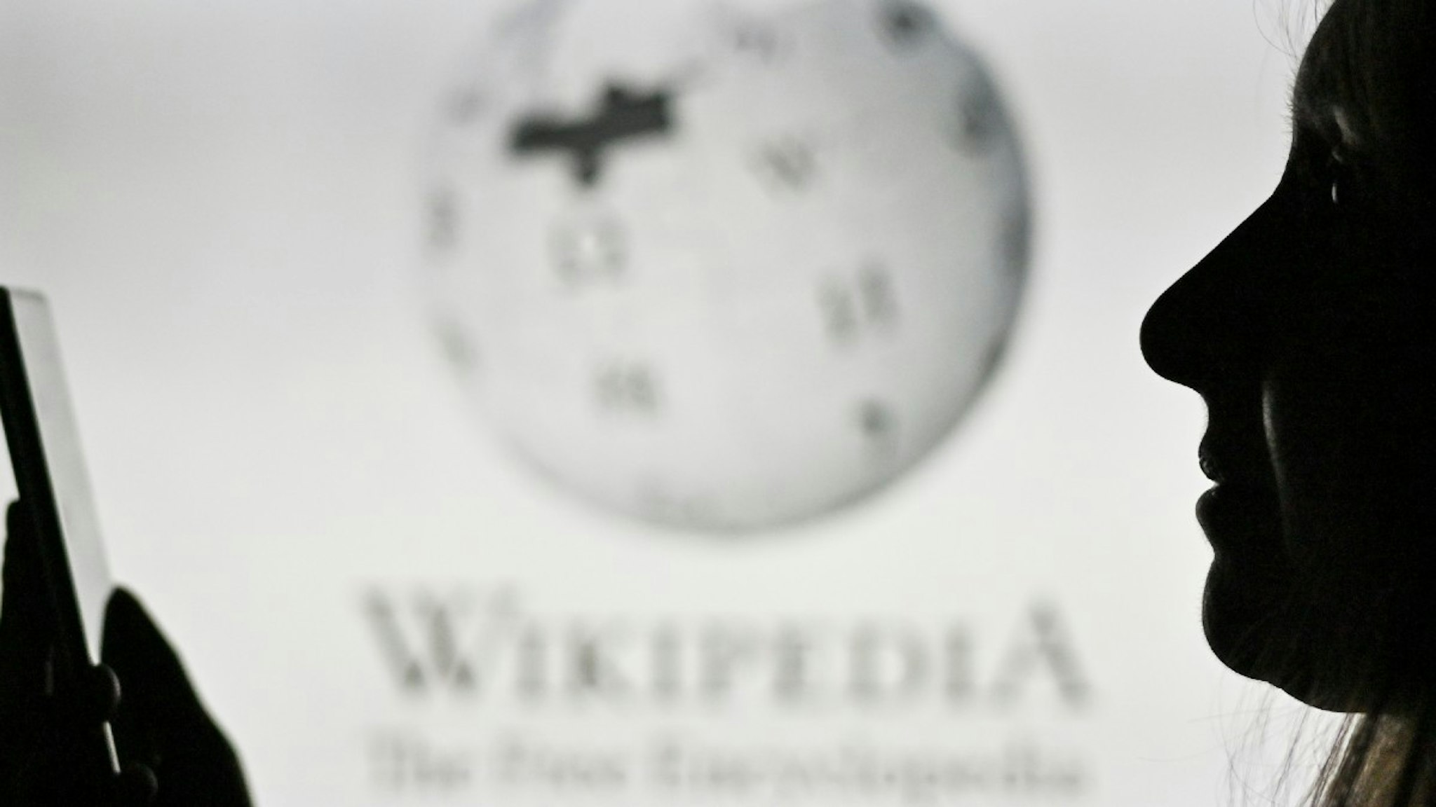 An image of a woman holding a cell phone in front of the Wikipedia logo displayed on a computer screen. On Tuesday, January 12, 2021, in Edmonton, Alberta, Canada.