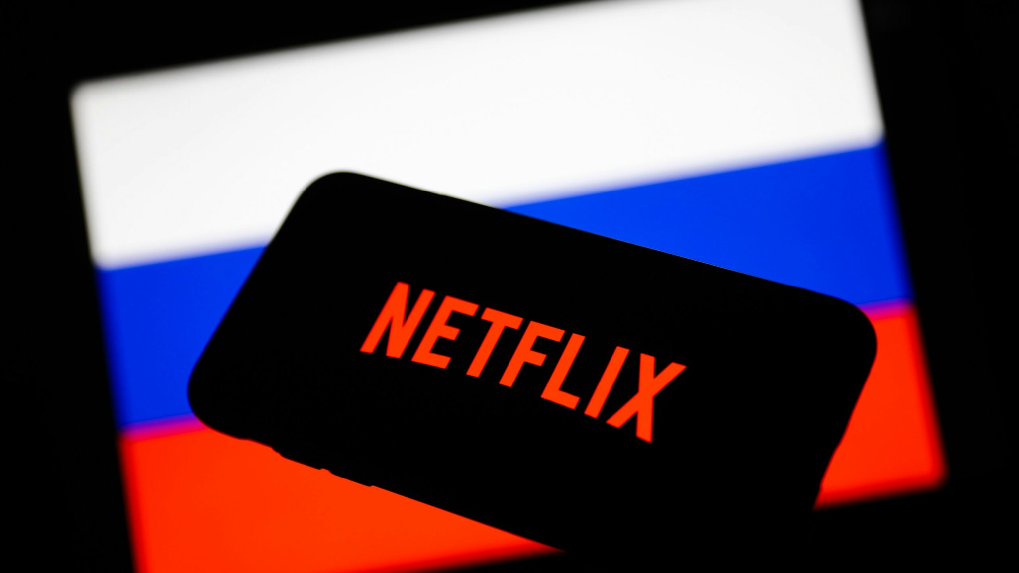 Netflix logo displayed on a phone screen and Russian flag displayed on a screen in the background are seen in this illustration photo taken in Krakow, Poland on March 1, 2022.