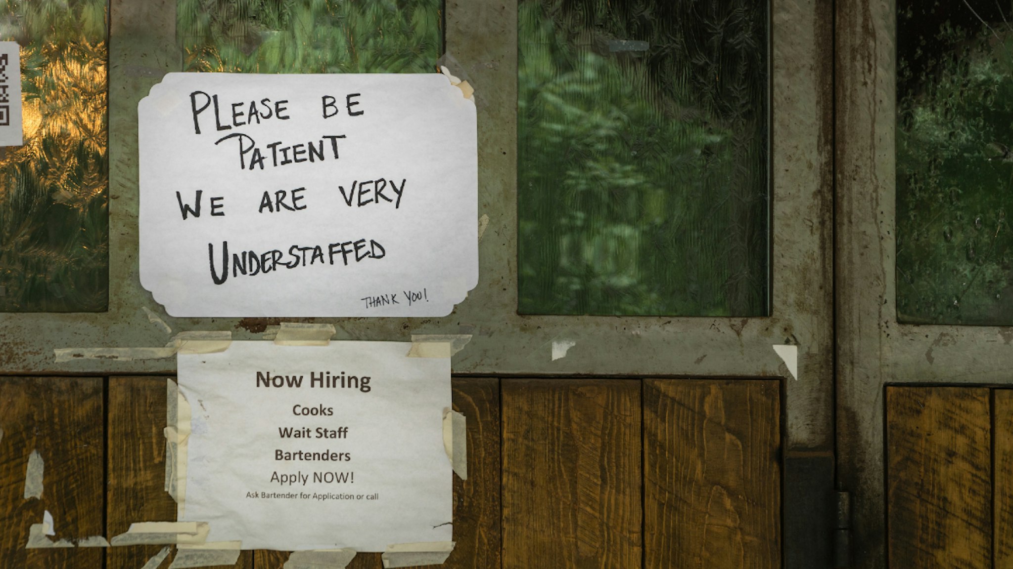 Rustic sign in restaurant window during time of Covid stating restaurant is understaffed and is hiring in a struggling economy, showing the plight of small businesses in the post-pandemic economy.