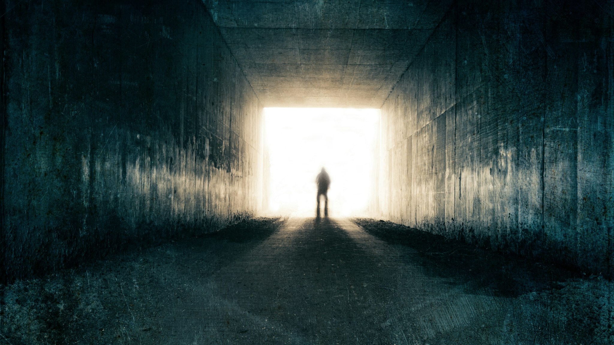 A silhouette of a blurred figure emerging from the light at the end of a dark sinister tunnel. With a grunge, vintage, grainy edit. - stock photo