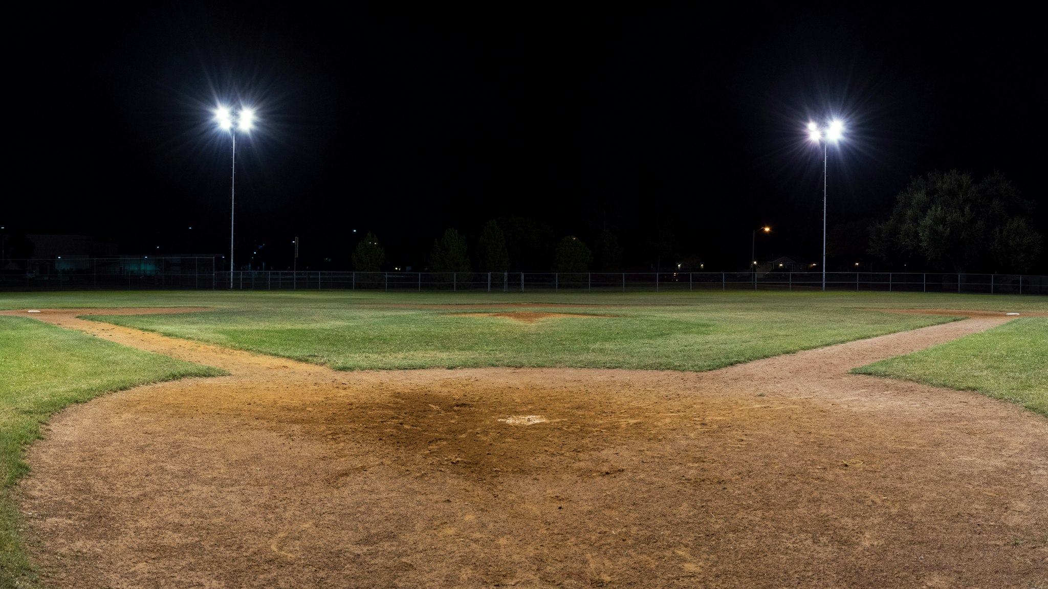 Panorama night photo of an empty baseball field at night with the lights on taken behind home plate and looking out across the pitcher's mound onto the field.