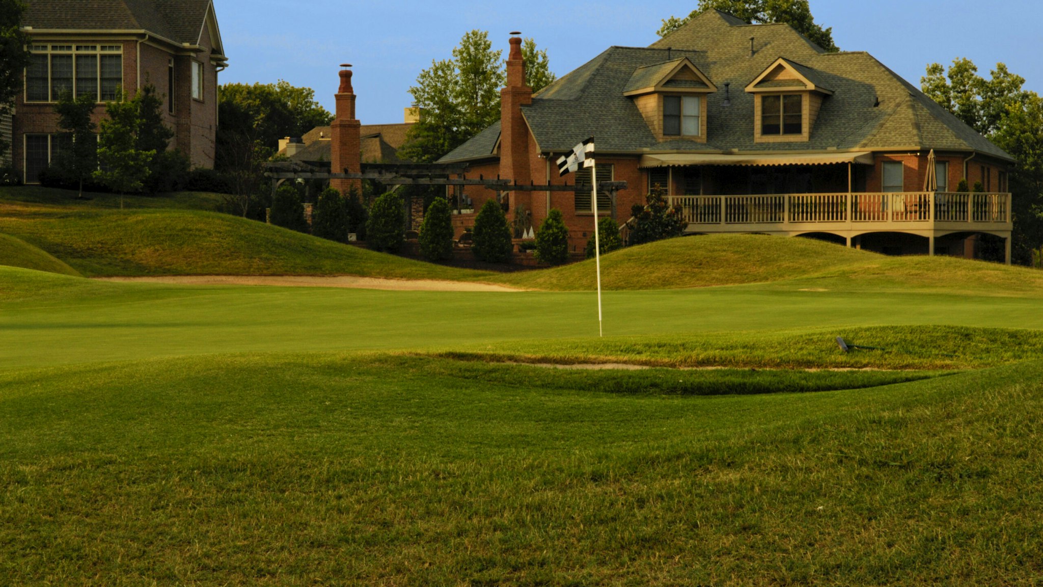 this color image is of a Putting Green on Professional Golf Course near Luxury Homes. the putting green has manicured grass or lawn with two sand traps around the outside of the putting green and putting hole. the grass is green and their are expensive houses or homes in the background. the photo was taken during the spring or summer. and the lighting is natural sunlight during the evening part of the day. there are also trees with leaves in the background.