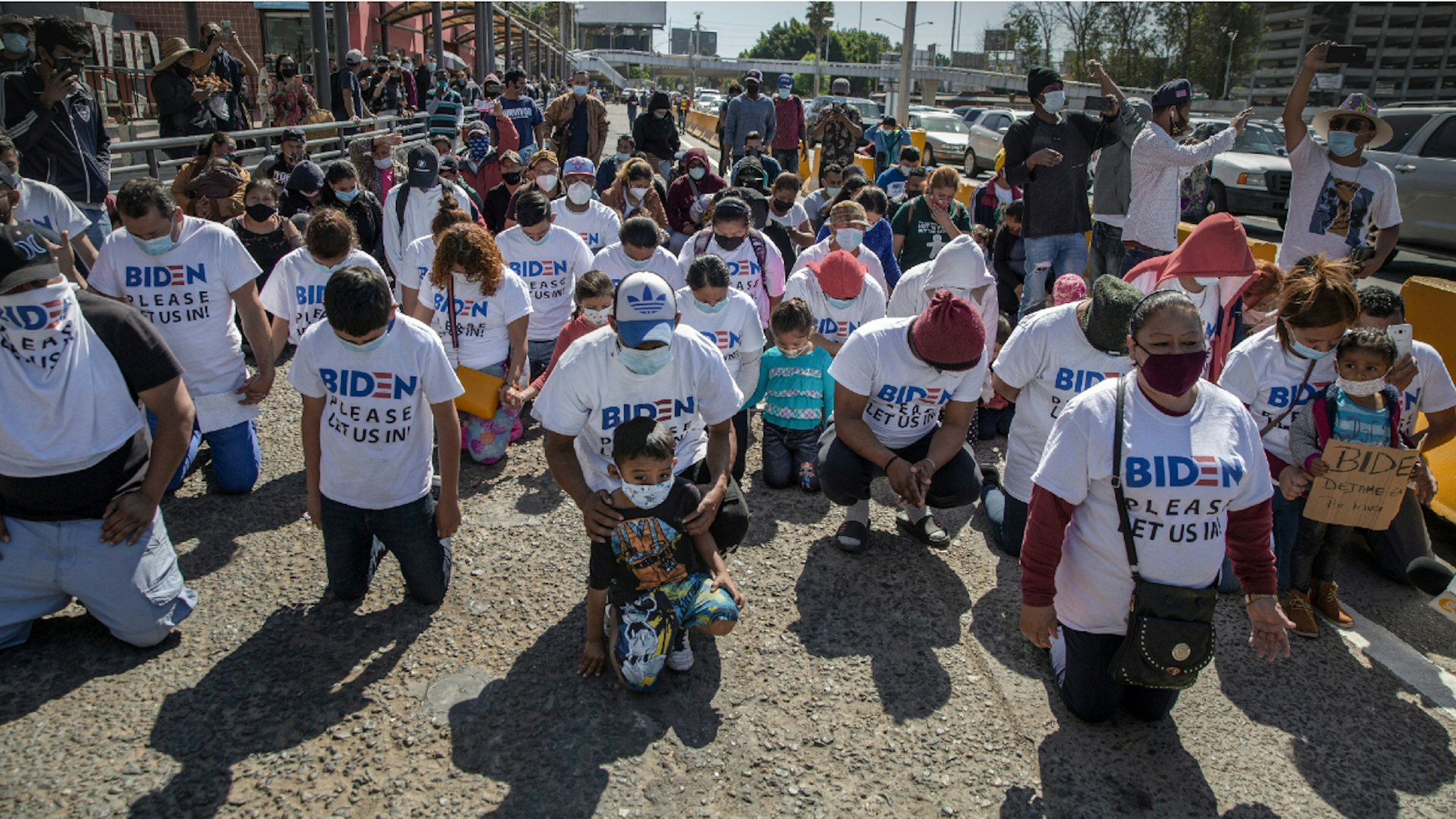 dpatop - 02 March 2021, Mexico, San Ysidro: A group of migrants wearing T-shirts that read "Biden, please let us in" kneel and pray at the border crossing.