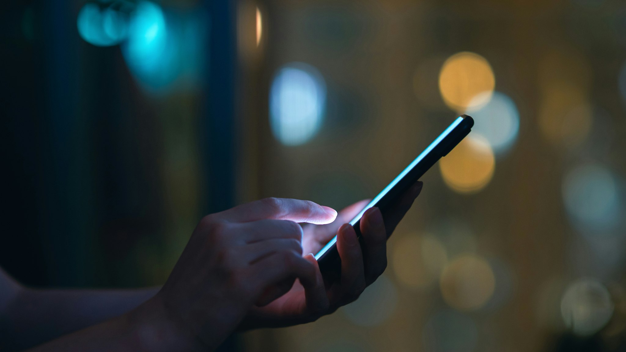 Close up of woman's hand using smartphone in the dark, against illuminated city light bokeh.