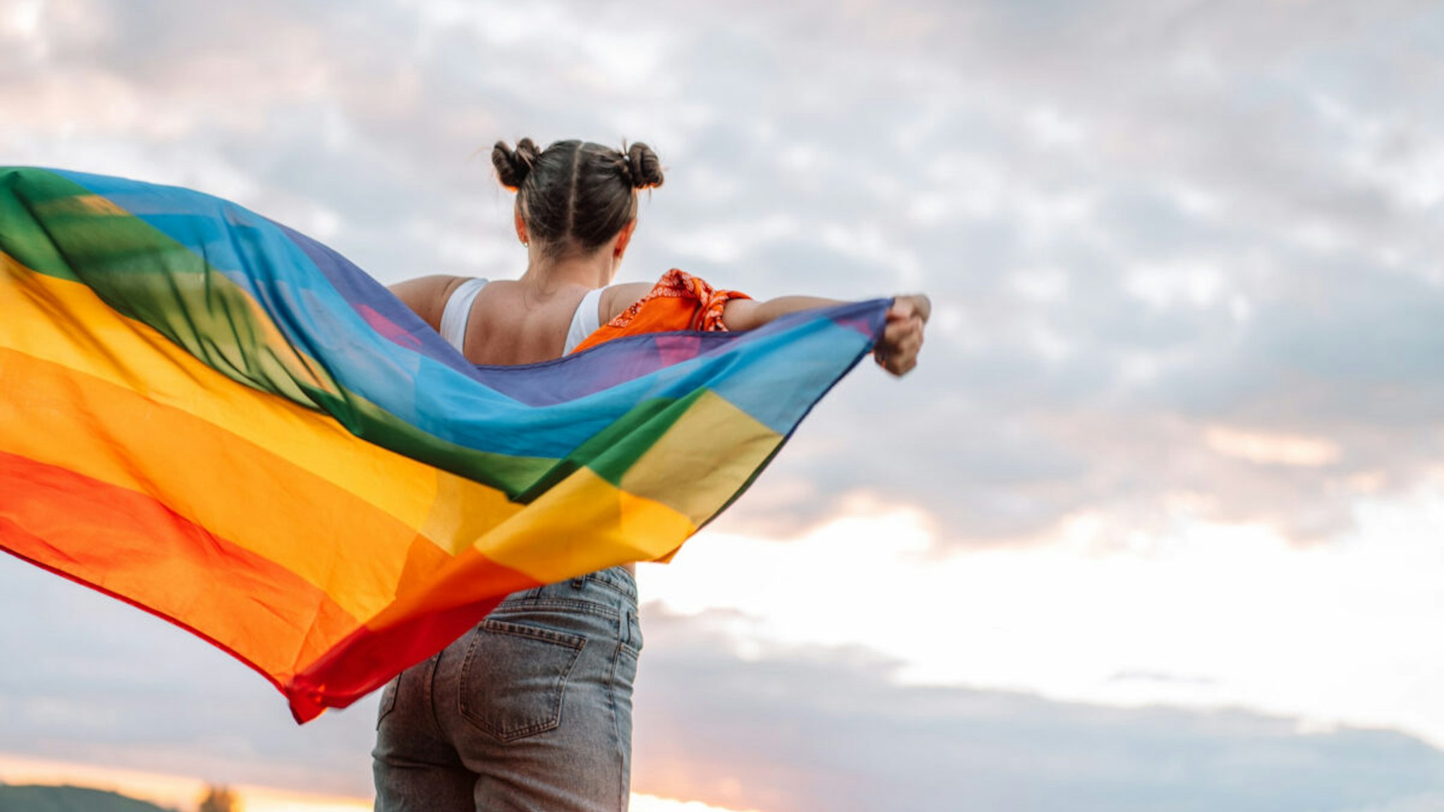 diversity Girl standing back and holding rainbow flag on sky background.