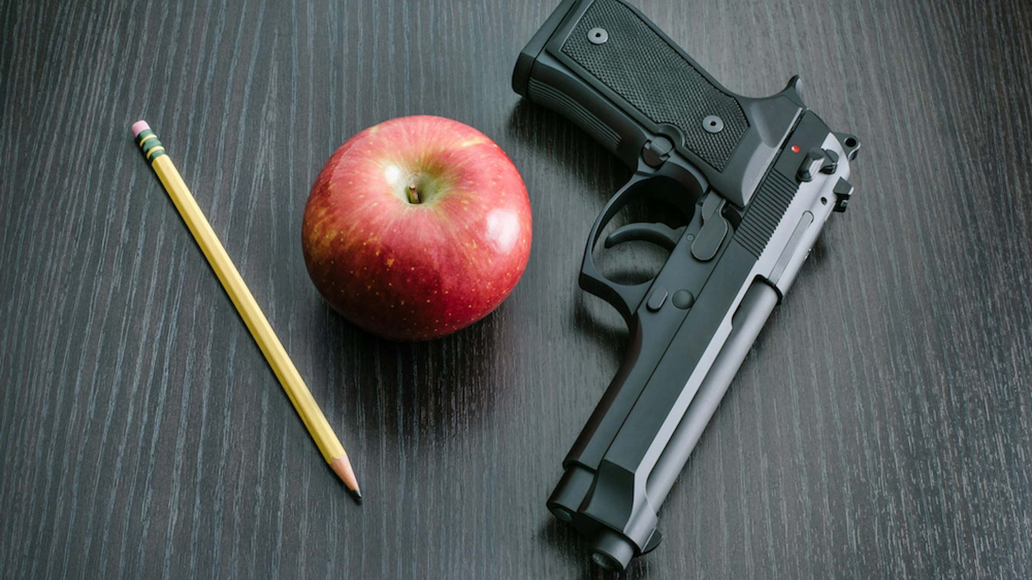 9mm Beretta 92FS type handgun with apple and pencil depicting the question whether to arm teachers in the classroom to defend students against active shooters. (kenlh/Getty Images)