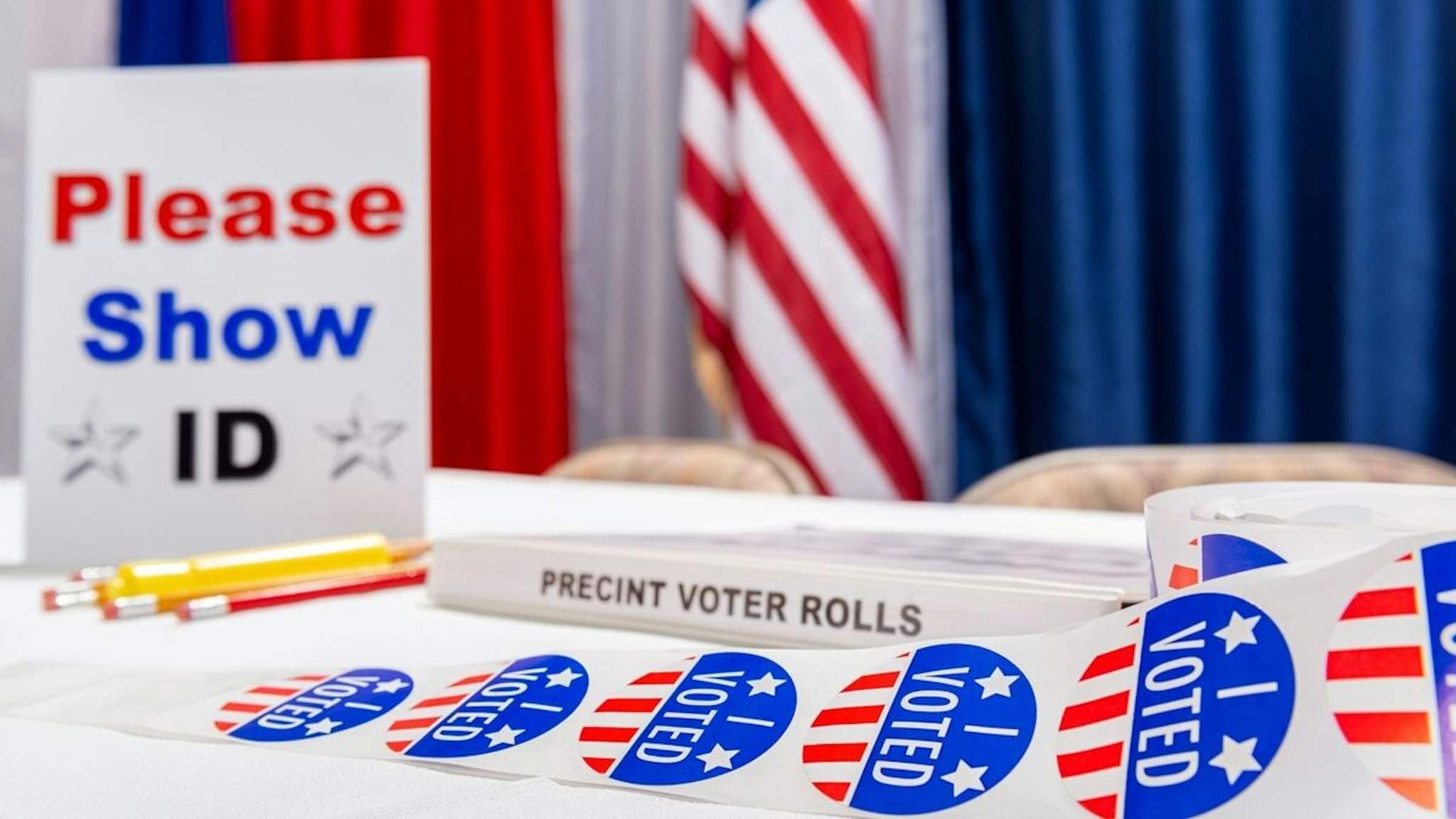 A roll of “I VOTED” stickers sitting on the table of an empty registration desk at a US polling station on election day with a “PLEASE SHOW ID” sign, red, white and blue bunting an American flag in the background.