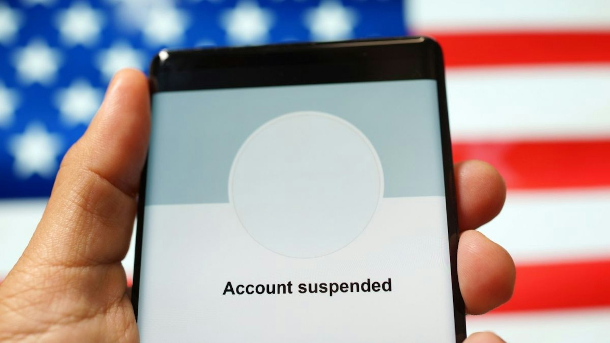 Close up an adult hand holding a smartphone showing "Account Suspended" from a social media application against the American flag background.