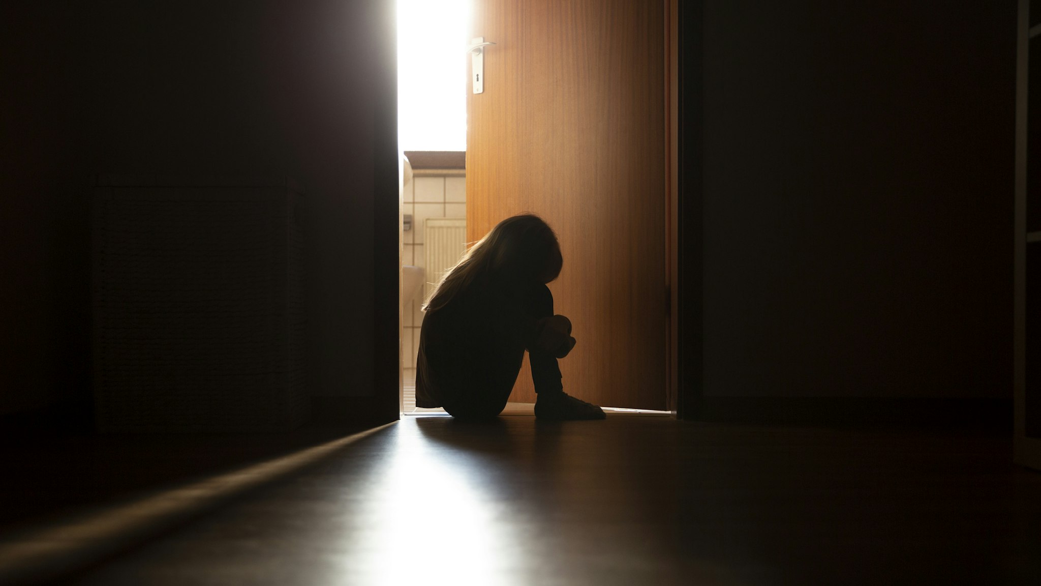 Despairing child sitting with head on knees in the dark frame of a doorway, backlit by a room behind flooded with daylight - stock photo