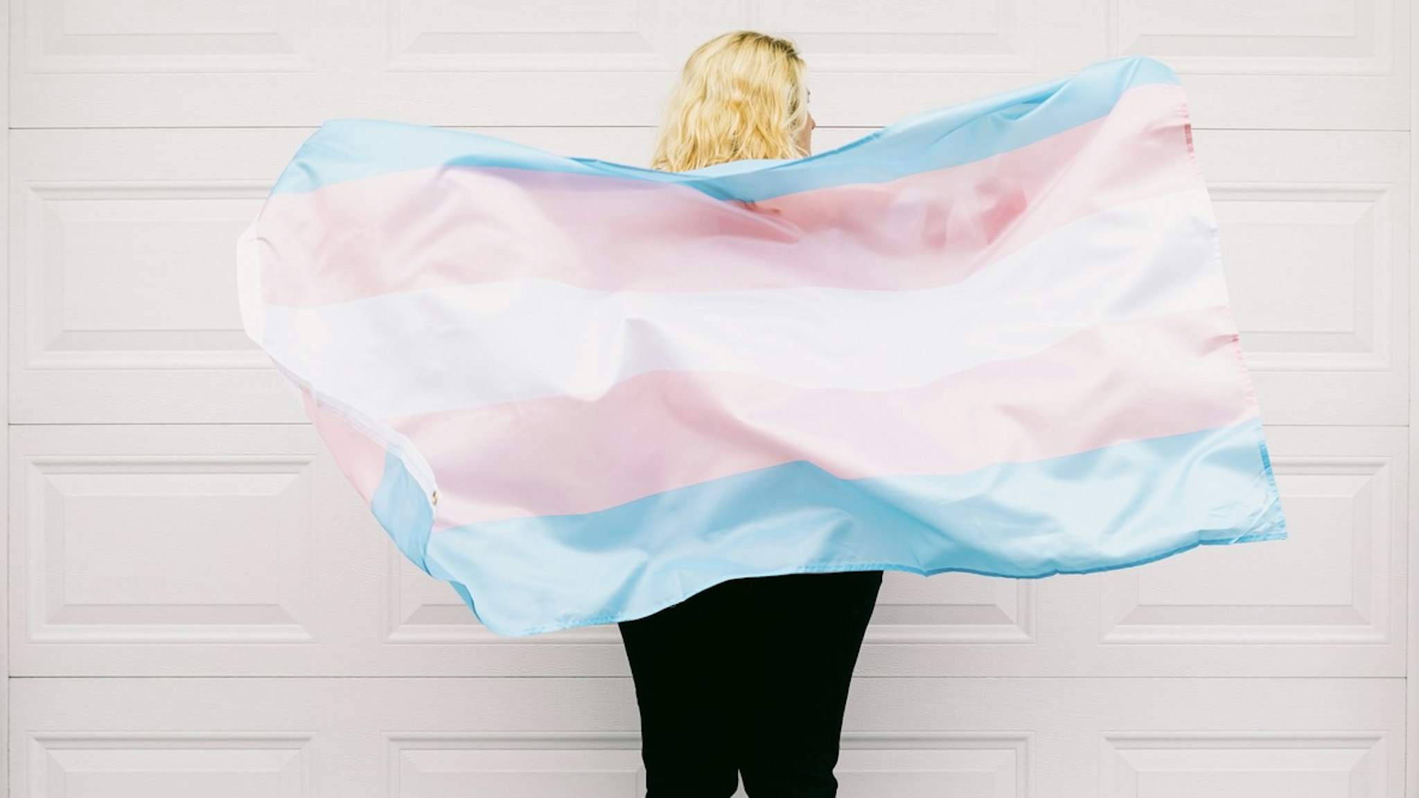 Transgender person from behind, wearing pink and white striped sweatshirt, holds transgender flag - stock photo Transgender person from behind, wearing pink and white striped sweatshirt, holds transgender flag DBenitostock via Getty Images