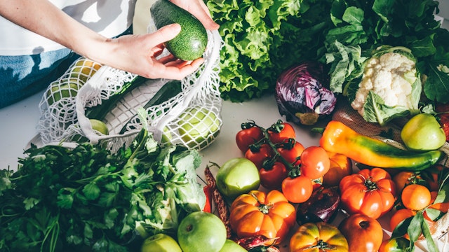 Young woman returned with purchases from grocery store takes fresh organic vegetables out of mesh bag putting on kitchen table at home close view