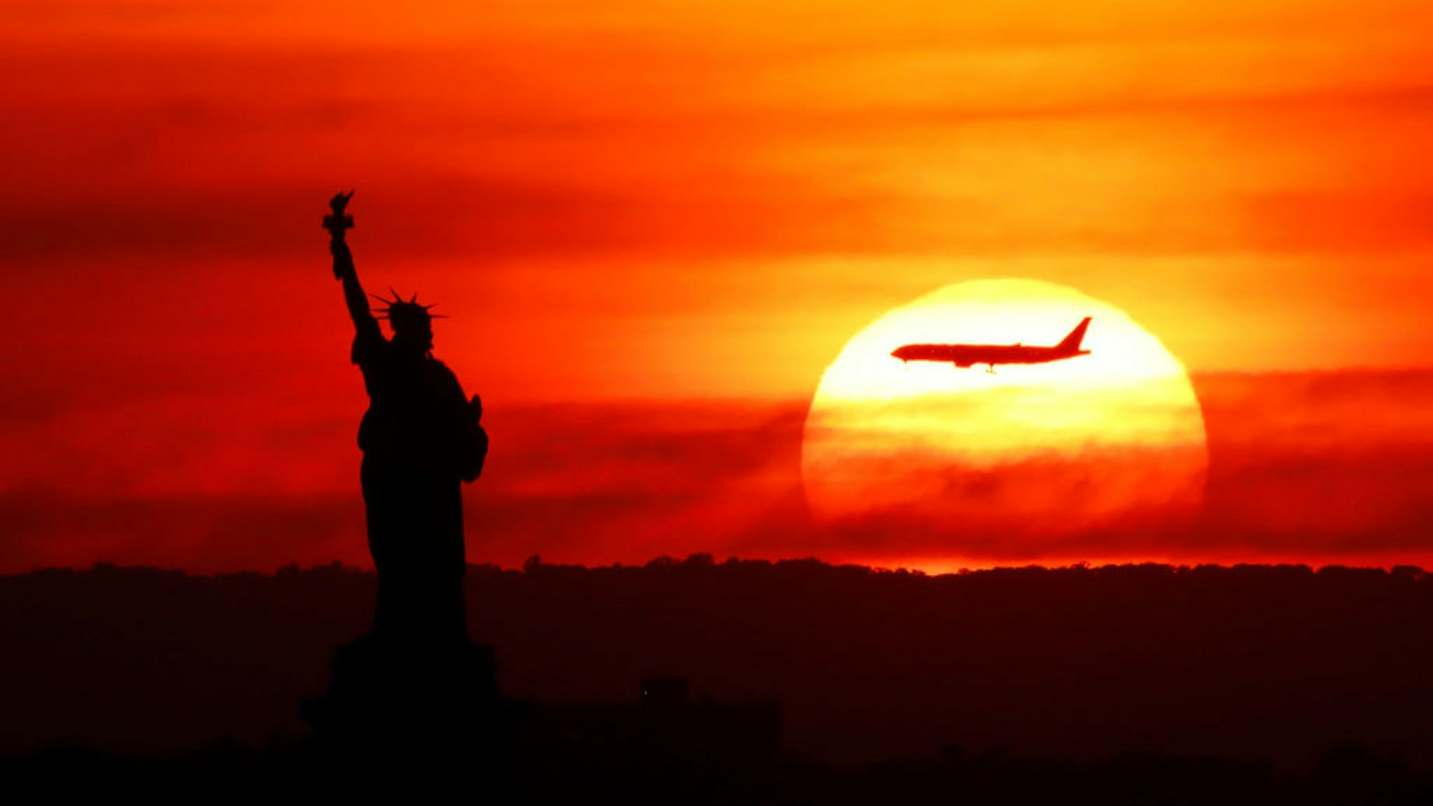 NEW YORK, NY - AUGUST 3: An airplane on approach to Newark Liberty Airport flies behind the Statute of Liberty as the sun sets on August 3, 2022, in New York City.