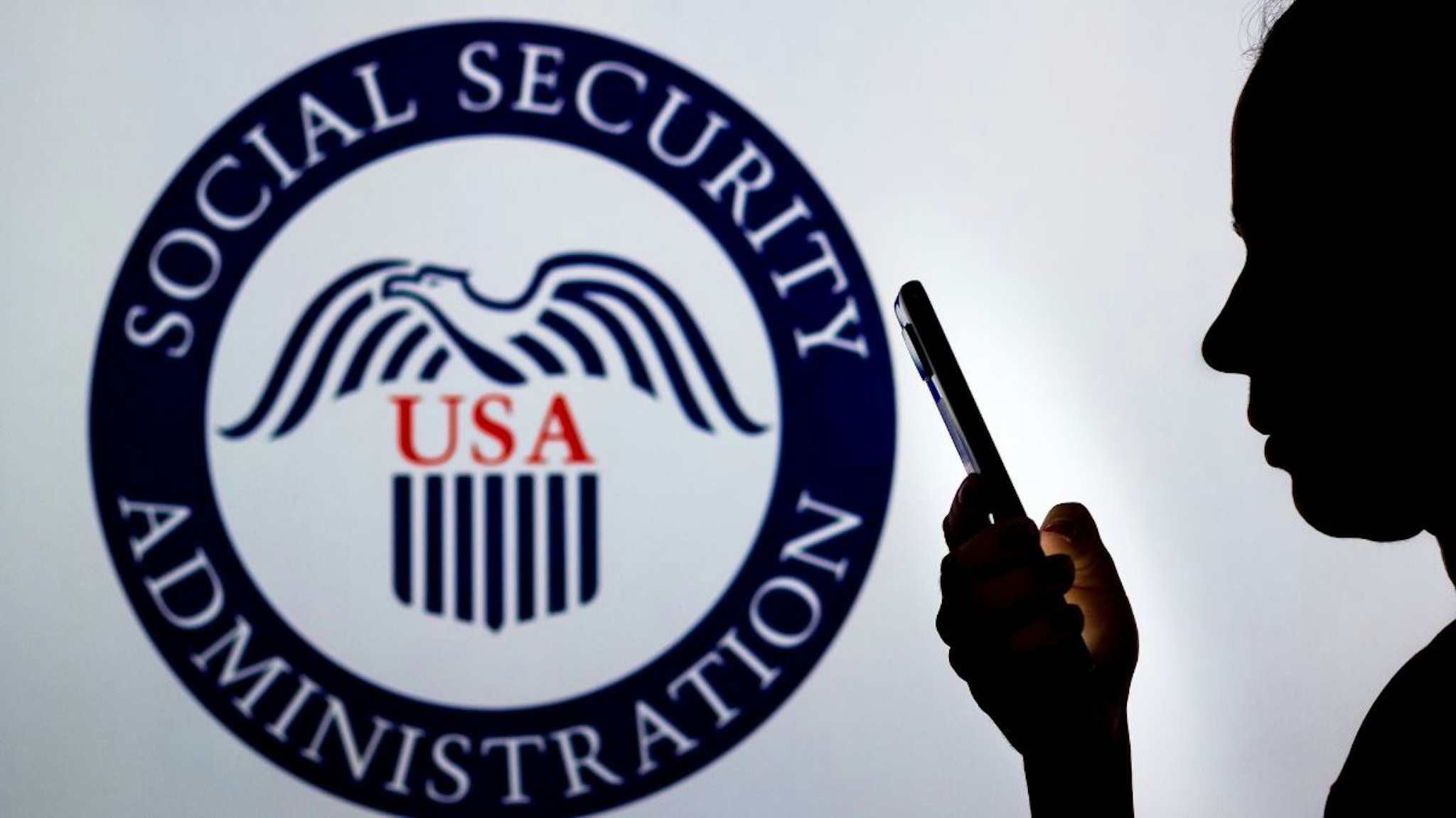 In this photo illustration, a woman's silhouette holds a smartphone with the United States Social Security Administration logo in the background.