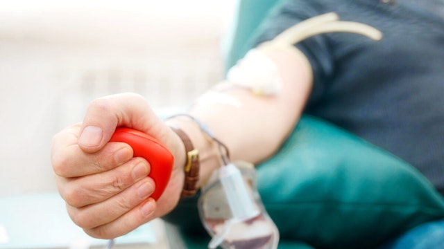 Topic of donation. Man donates blood in hospital. Man's hand squeezes rubber heart. Close-up. Donor sits in chair. Background.