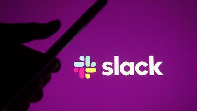 UKRAINE - 2021/02/21: In this photo illustration a silhouette hand holding a smartphone is seen in front of Slack logo of a business communication platform.