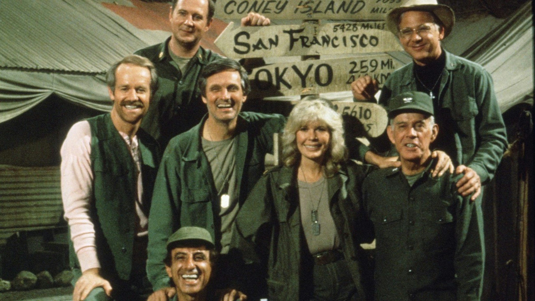 Portrait the cast of the television show 'MASH,' mid- to late-1970s, From left American actors Mike Farrell (with light sleeves), David Ogden Stiers (in rear), Alan Alda, Jamie Farr (sitting in foreground), Loretta Swit, William Christopher (in rear), and Harry Morgan