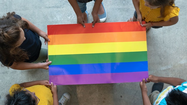 A group of multi-ethnic elementary students stand around a colorful gay pride poster.
