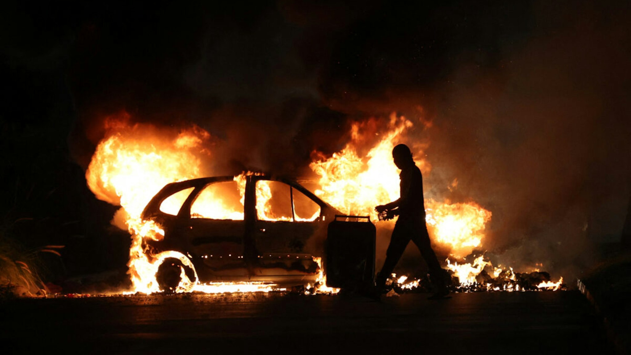 TOPSHOT - A protester walks by a burning car during clashes with police in Le Port, French Indian Ocean island of La Reunion, on June 30, 2023, three days after a 17-year-old boy was shot in the chest by police at point-blank range in Nanterre, a western suburb of Paris. A third consecutive night of violence in France sparked by the killing of a teenager by a policeman during a traffic stop has left 249 police and gendarmes injured, the interior ministry announced on June 30, 2023.