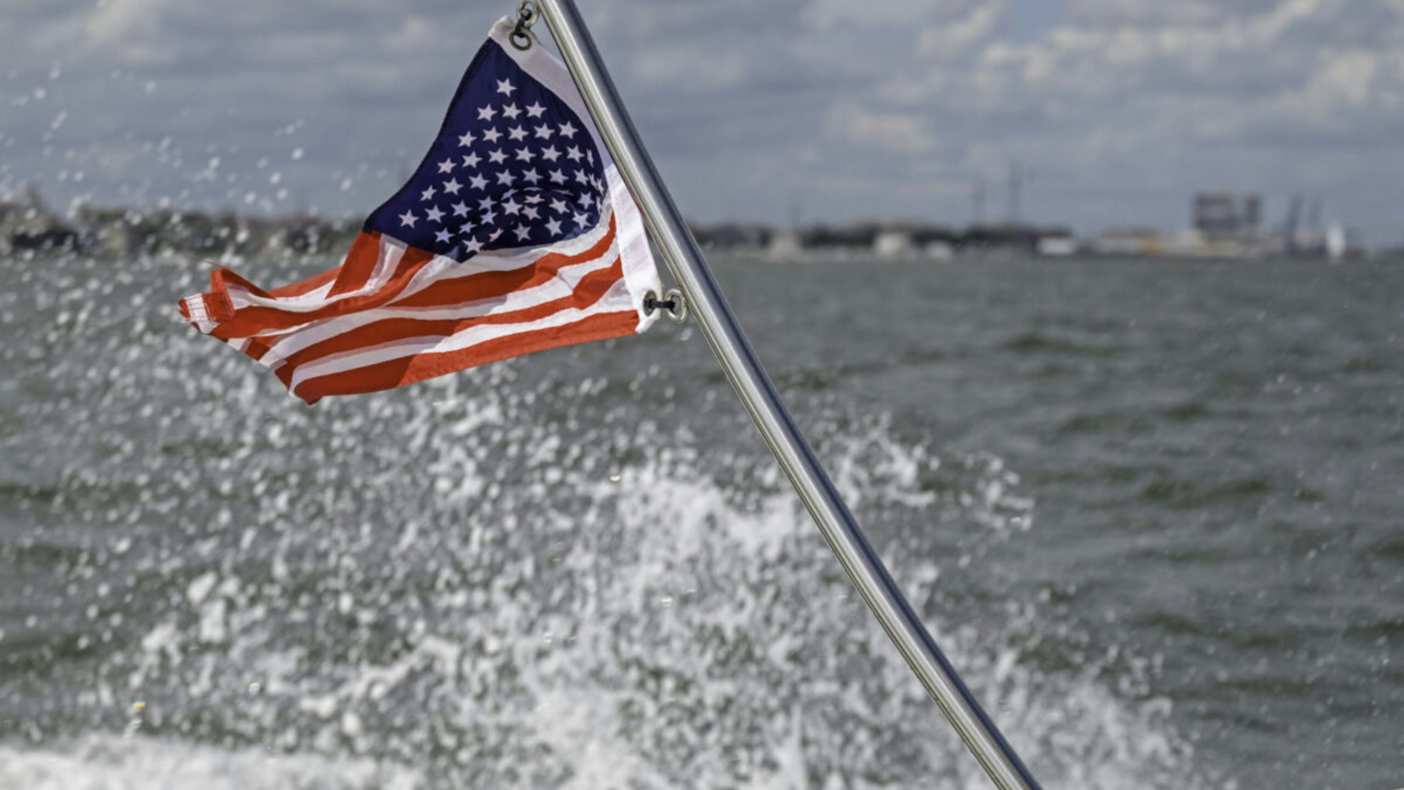 American flag flapping in the wind at the stern of a boat, showing water splashing and coastline in the background.