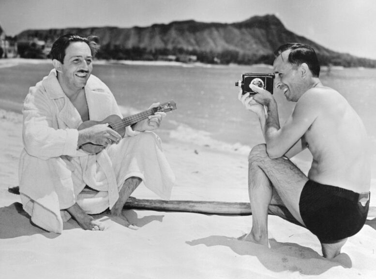 (Original Caption) Walt Disney, celebrated cartoonist and the creator of Mickey Mouse, is shown on the beach at Waikiki playing on a ukulele, while his brother and business manager, Roy, makes him the subject of a movie. Bettmann/Contributor. Getty Images.