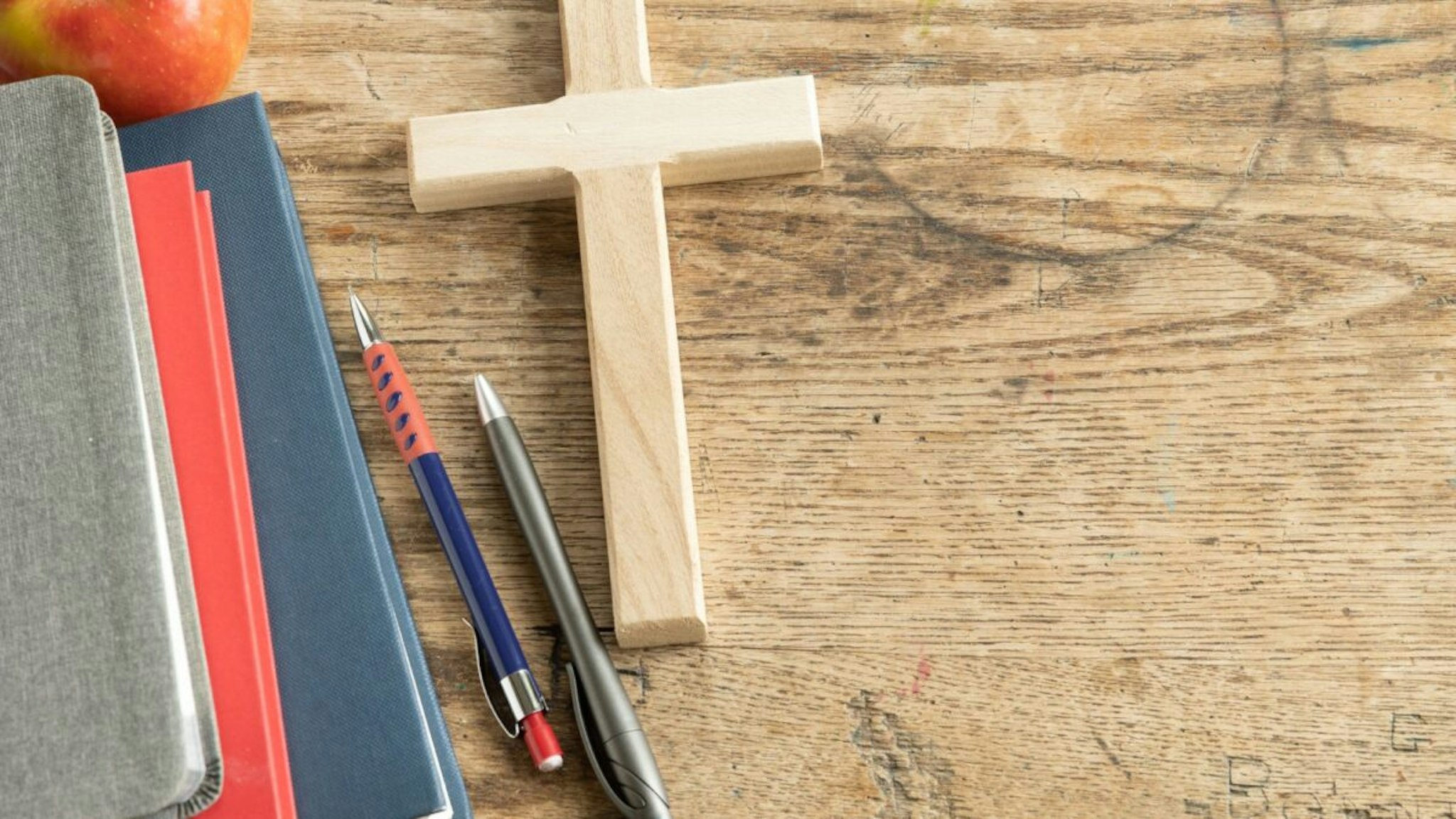 Wood Christian cross laying on a vintage wood desk with a stack of grey, red and navy blue books, a red apple and a pen and pencil, shot from above with copy space