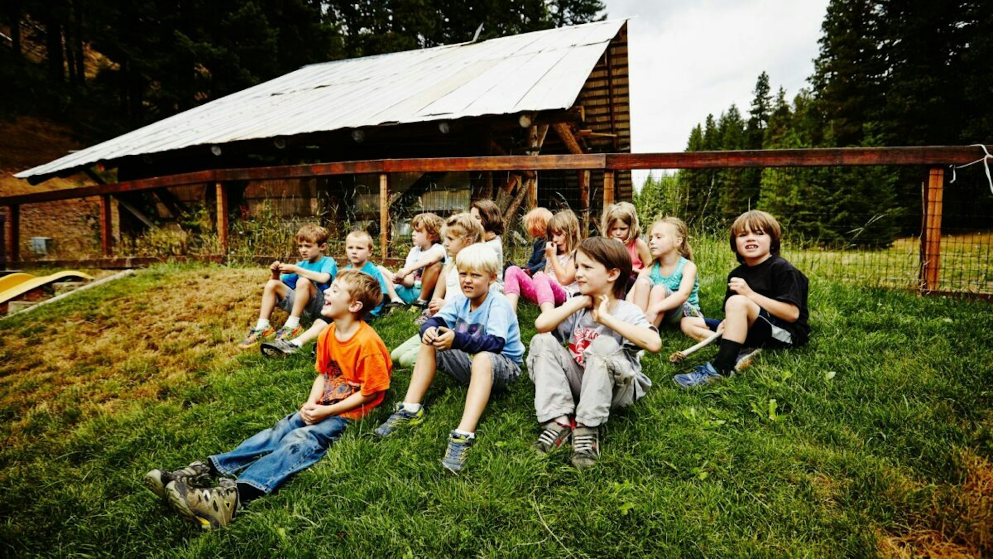 Group of young kids sitting on grass hillside of playground waiting for camp counselor at summer camp