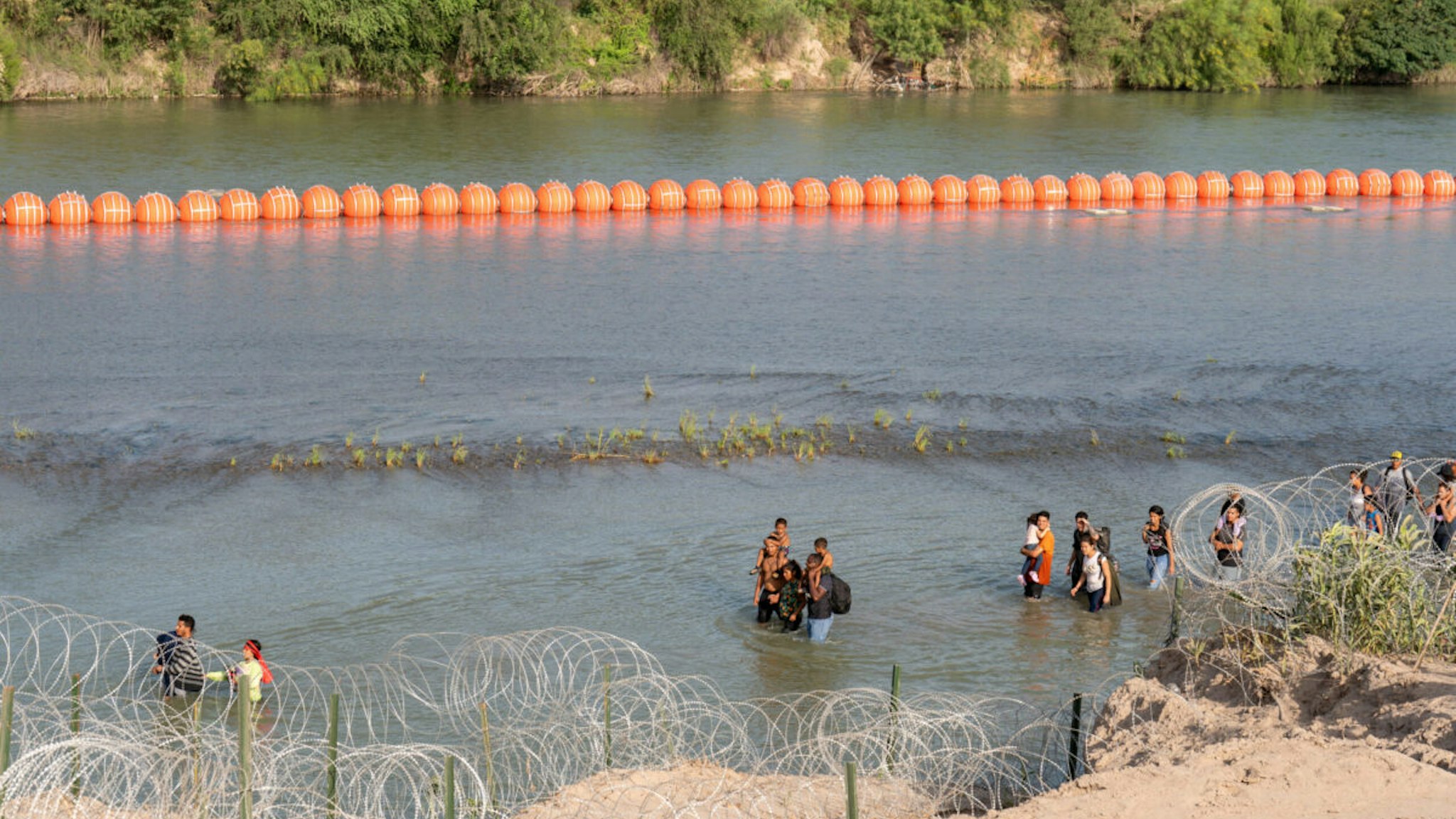TOPSHOT - Migrants walk by a string of buoys placed on the water along the Rio Grande border with Mexico in Eagle Pass, Texas, on July 16, 2023. The buoy installation is part of an operation Texas is pursuing to secure its borders, but activists and some legislators say Governor Greg Abbott is exceeding his authority.